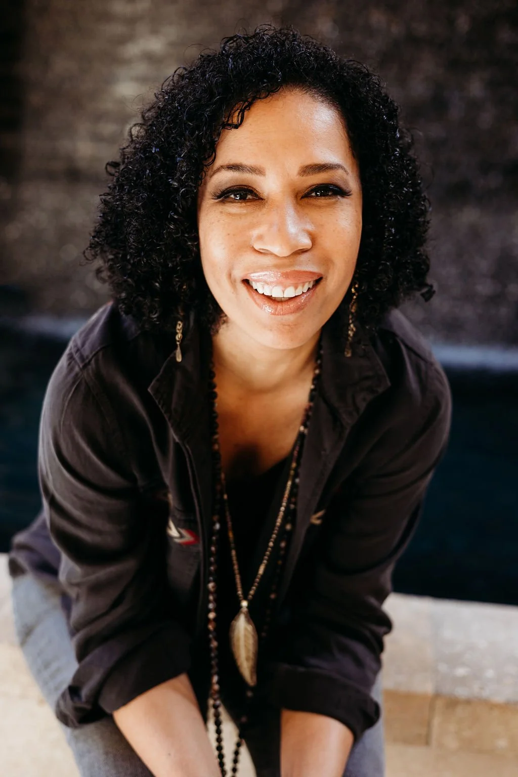 A woman with curly black hair smiling, wearing a black jacket with rolled-up sleeves, a beaded necklace with a large pendant, and earrings, against a dark background.