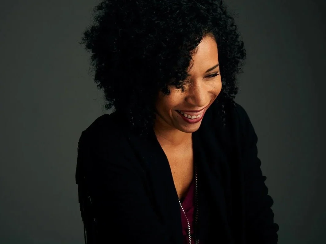 A woman with curly black hair smiling and looking down, wearing a black top with a darker background.