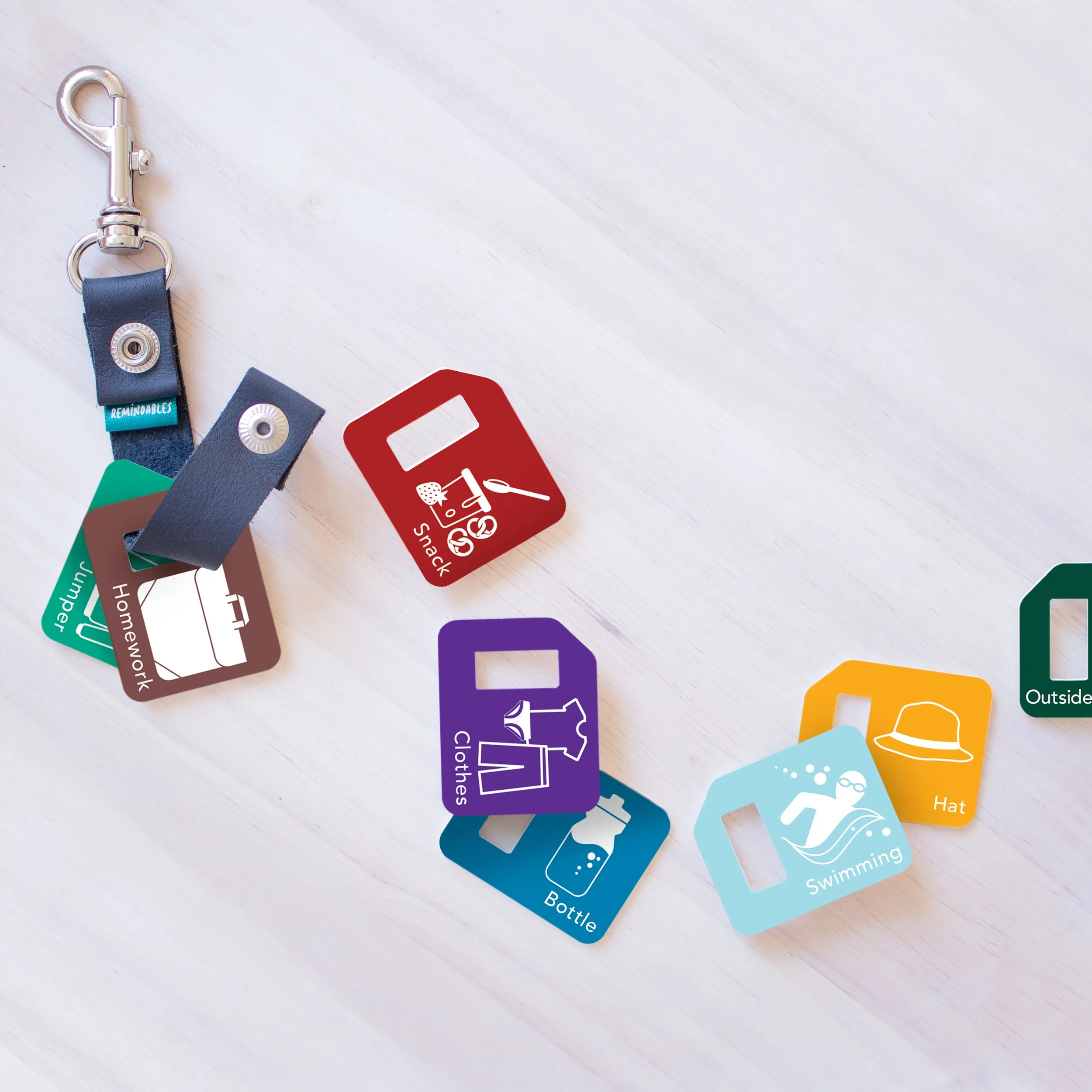 Colored reminder tags with various icons and labels such as snack, homework, clothes, and swimming, attached to a key clip on a white surface.