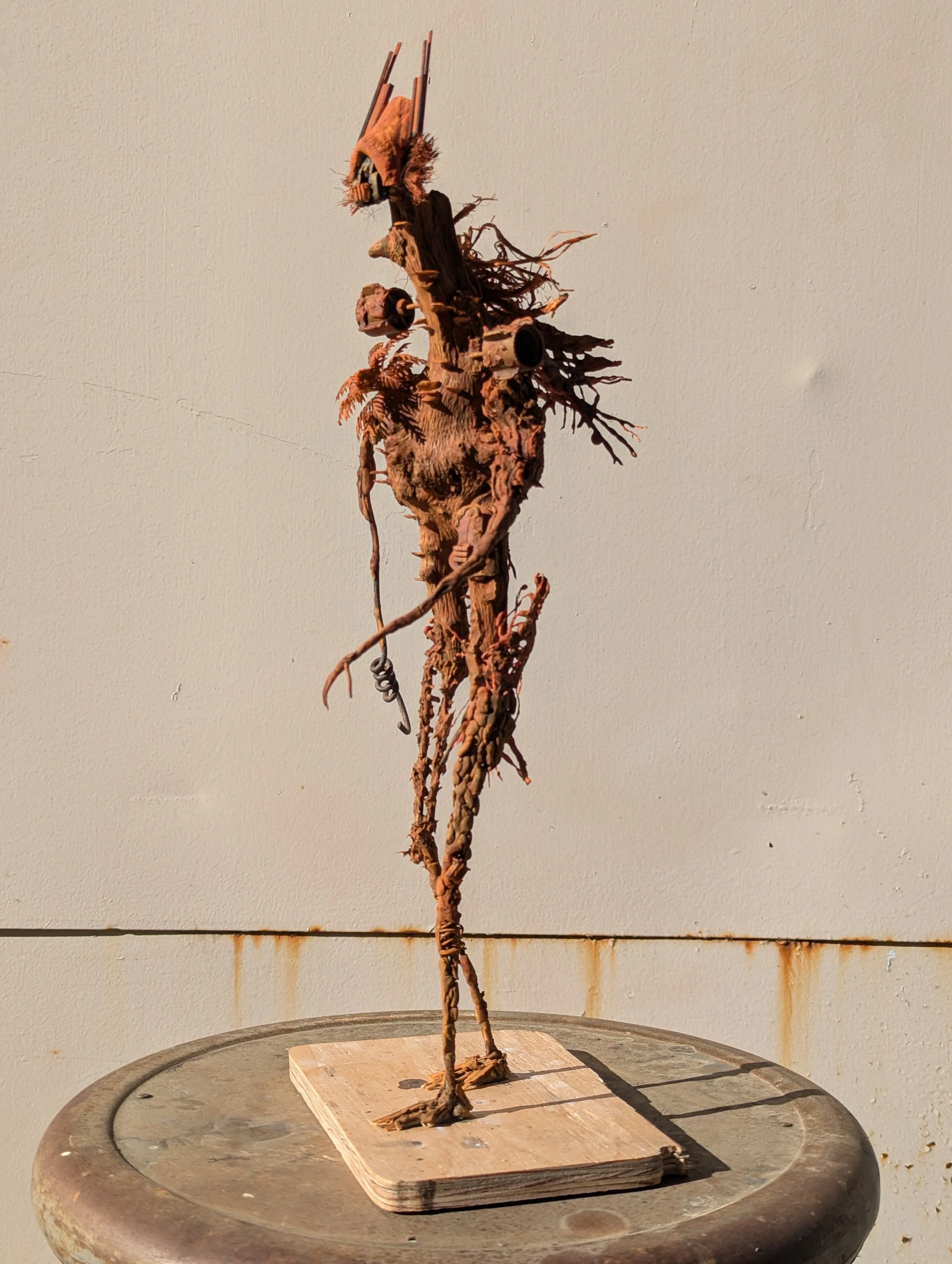 An abstract sculpture made of intertwined natural materials, resembling a tall, humanoid figure, on a wooden base against a plain wall.