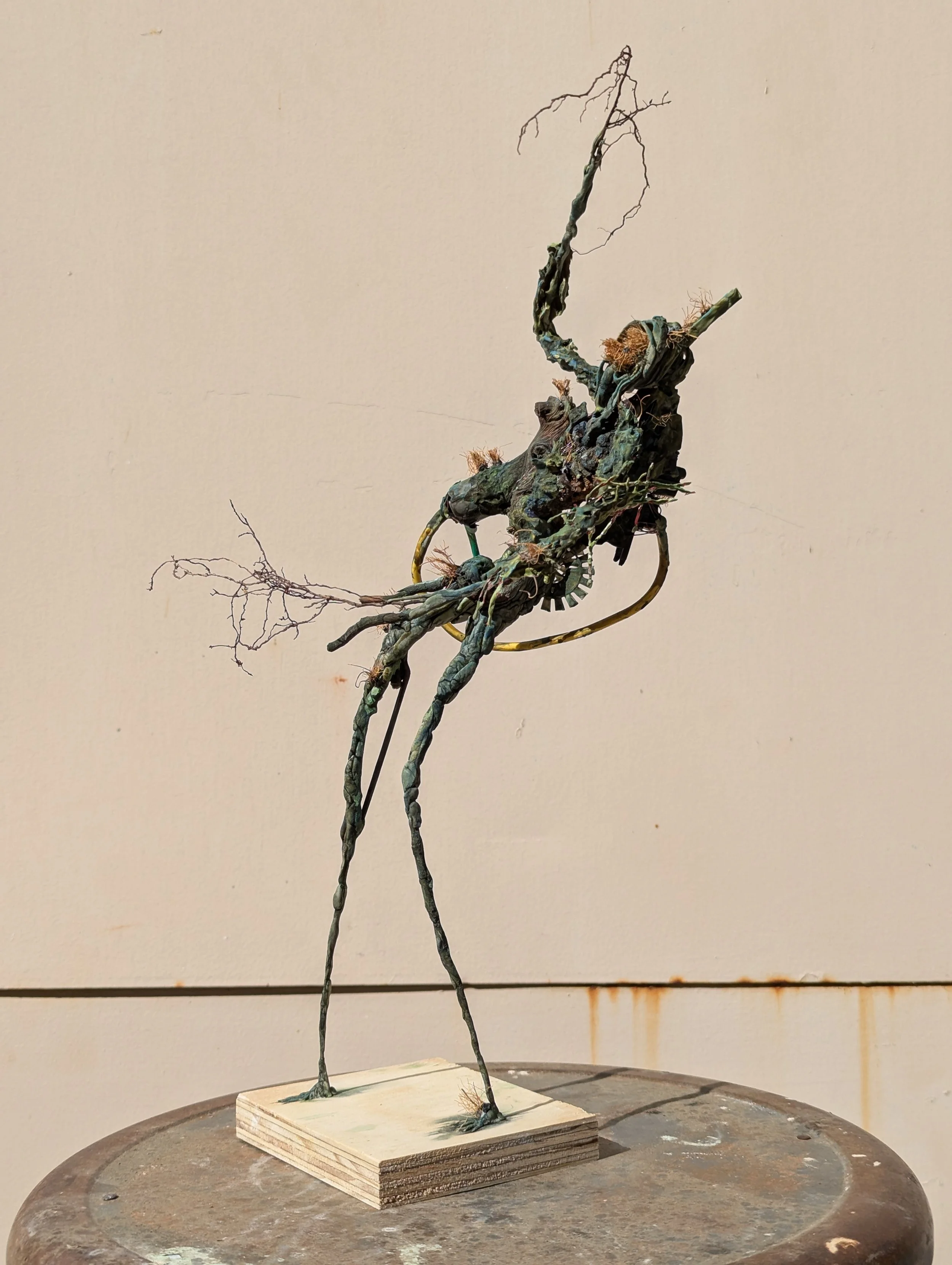 An abstract sculpture resembling a humanoid figure with elongated legs and arms, made of various found objects and wire, standing on a wooden base against a plain background.