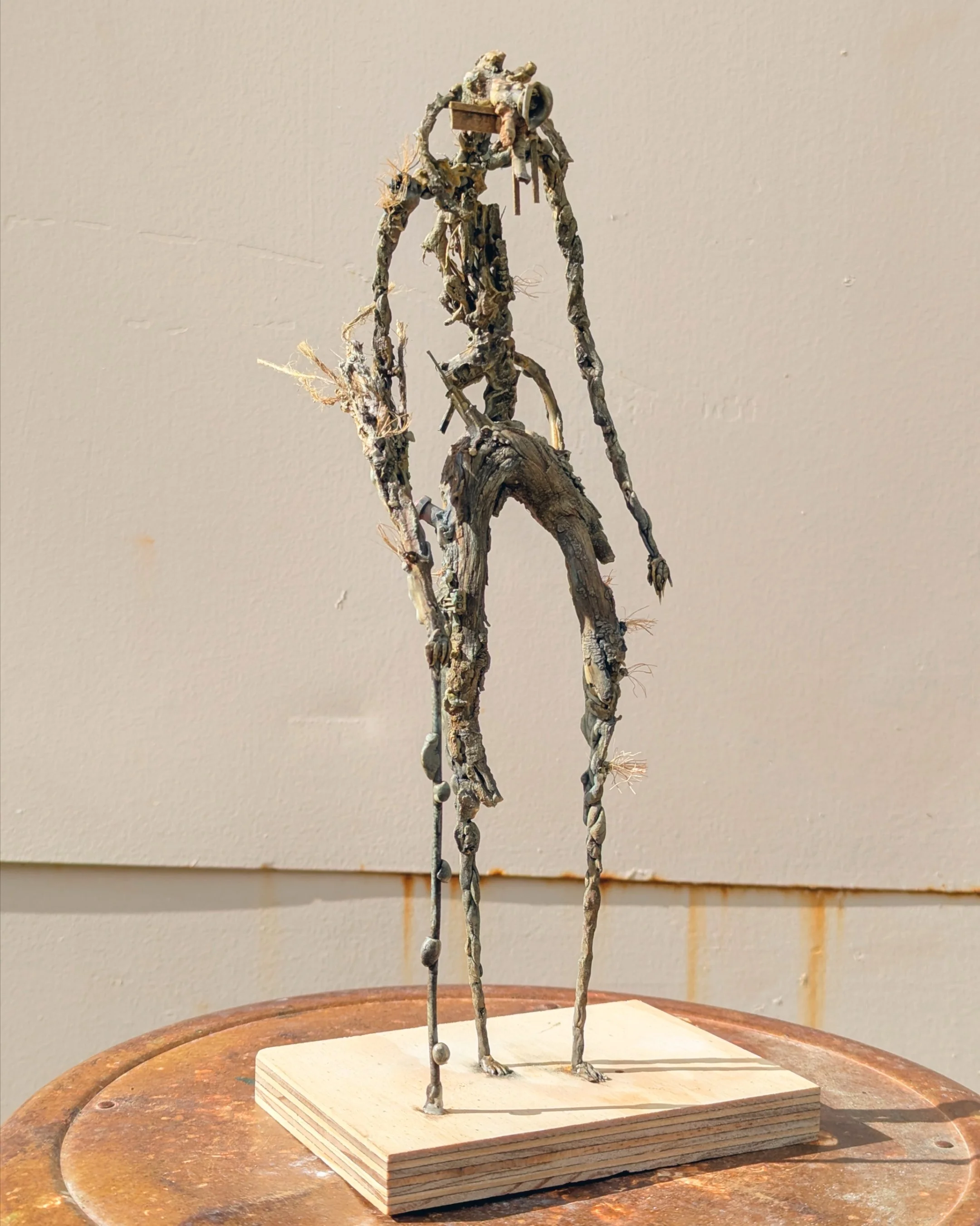 A tall, abstract sculpture of a human figure made of metal and found objects, standing on a wooden base against a plain wall.