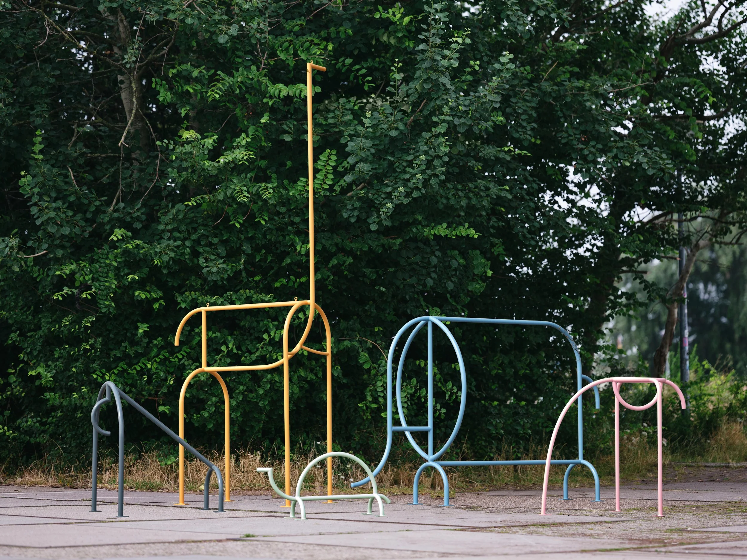 Colorful abstract outdoor sculpture resembling various playground equipment shapes, set against a background of green trees.