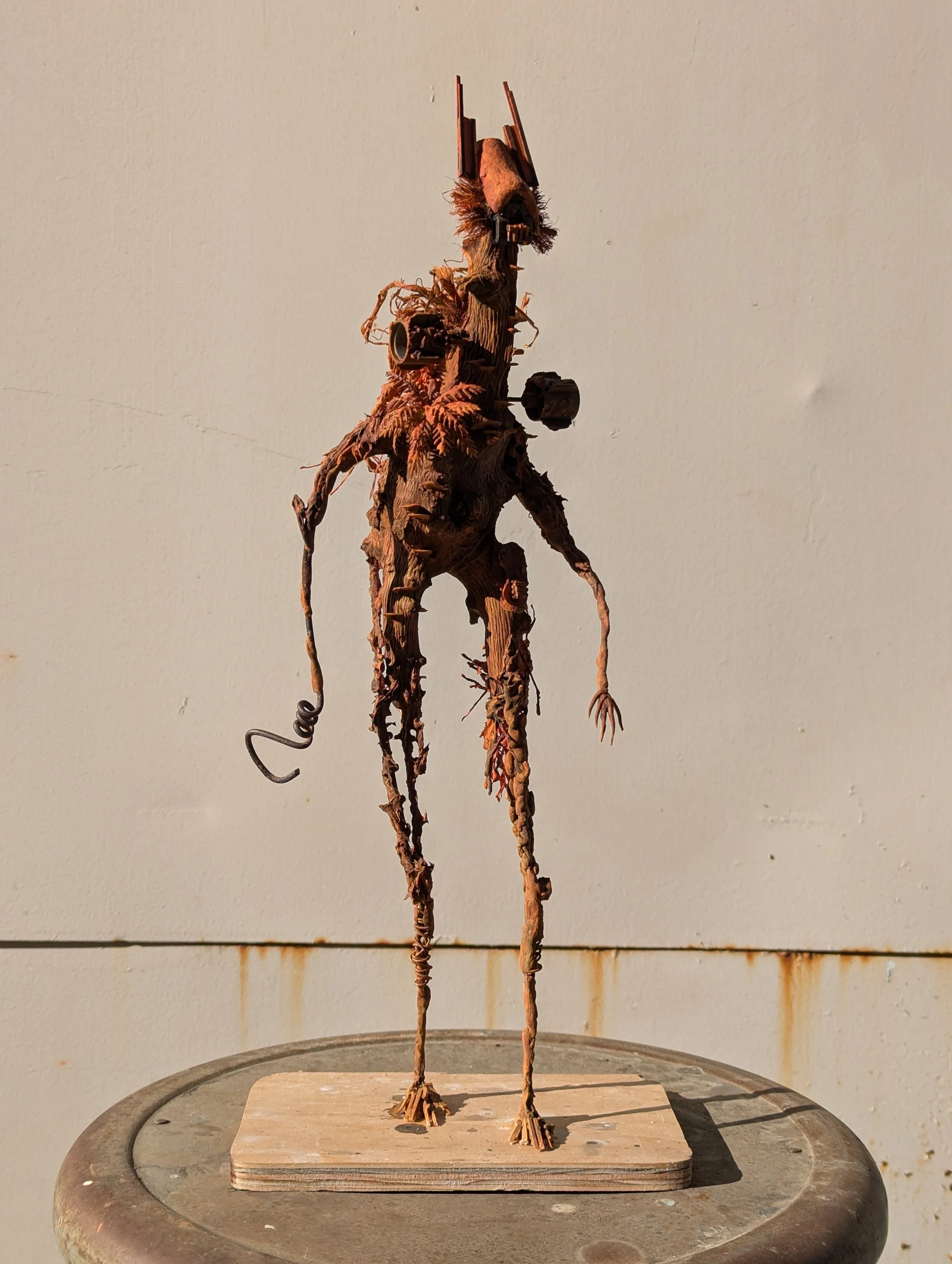 A tall humanoid sculpture made from twigs, with added objects like cups and wood pieces for details, standing on a wooden base.