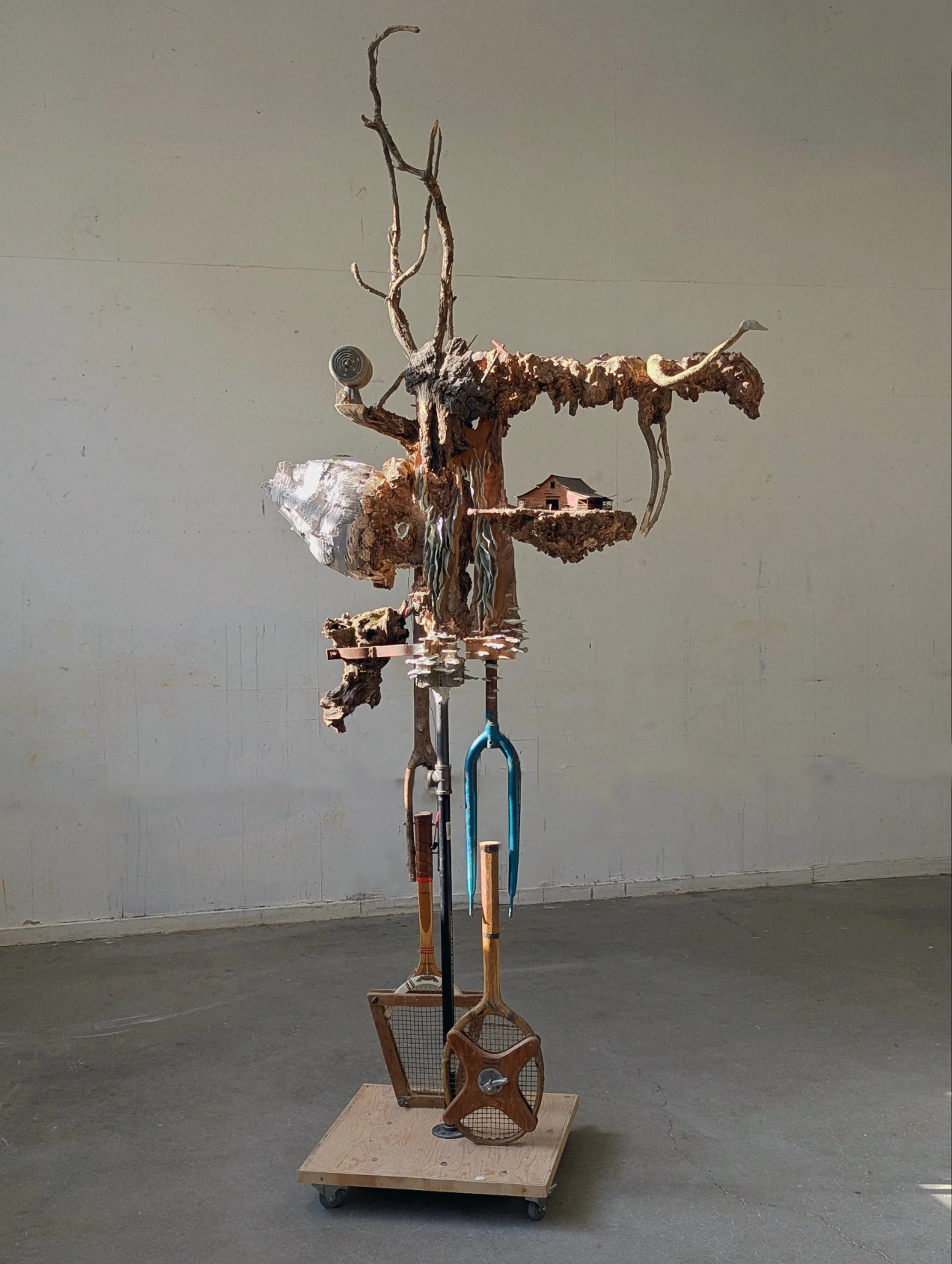 Mixed media sculpture of a stylized tree with various objects attached, mounted on a wheeled wooden platform in a gallery space.