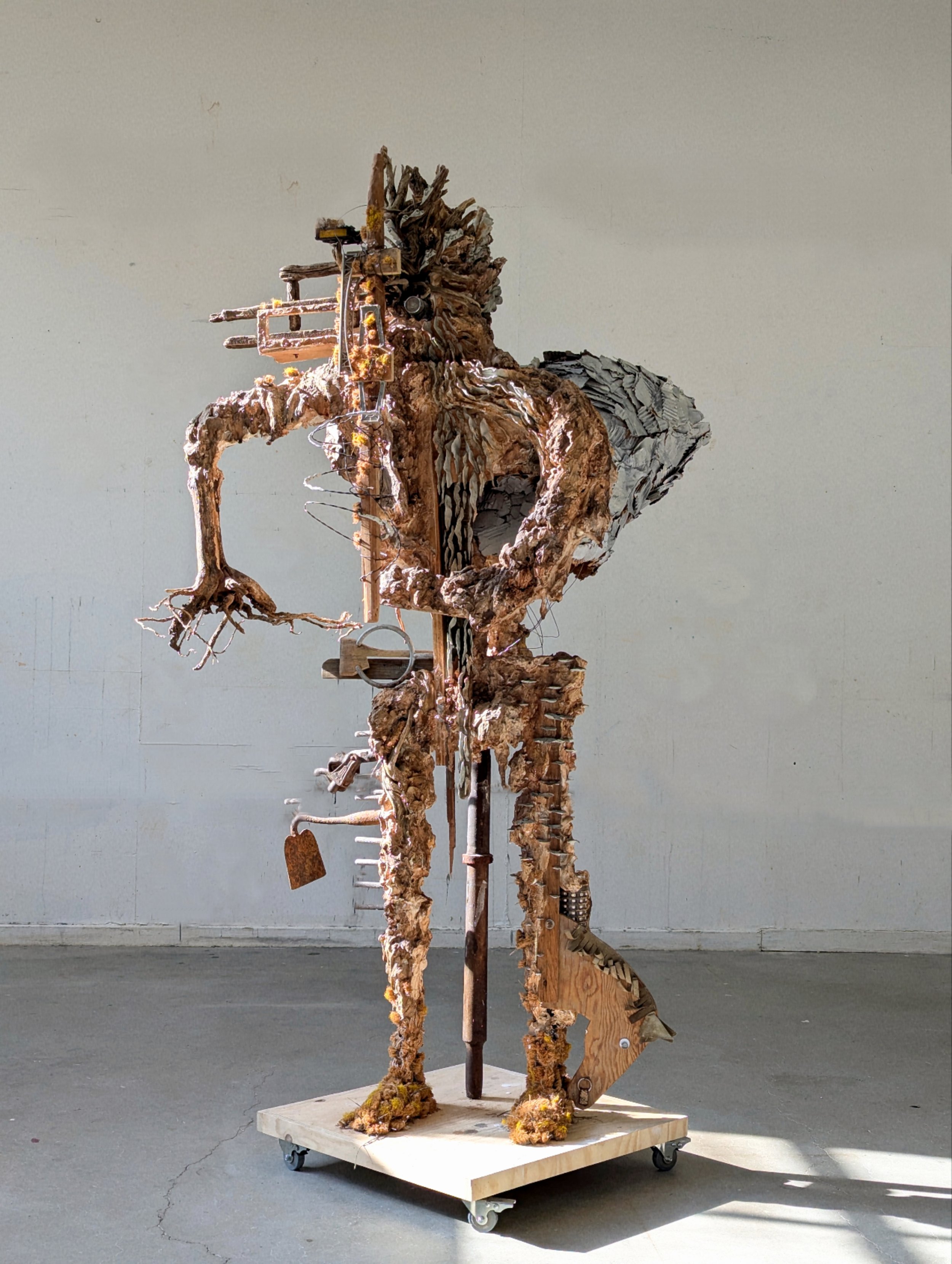 Abstract sculpture resembling a humanoid figure made from driftwood, metal, and other found objects, mounted on a wooden platform with wheels, set against a plain wall.
