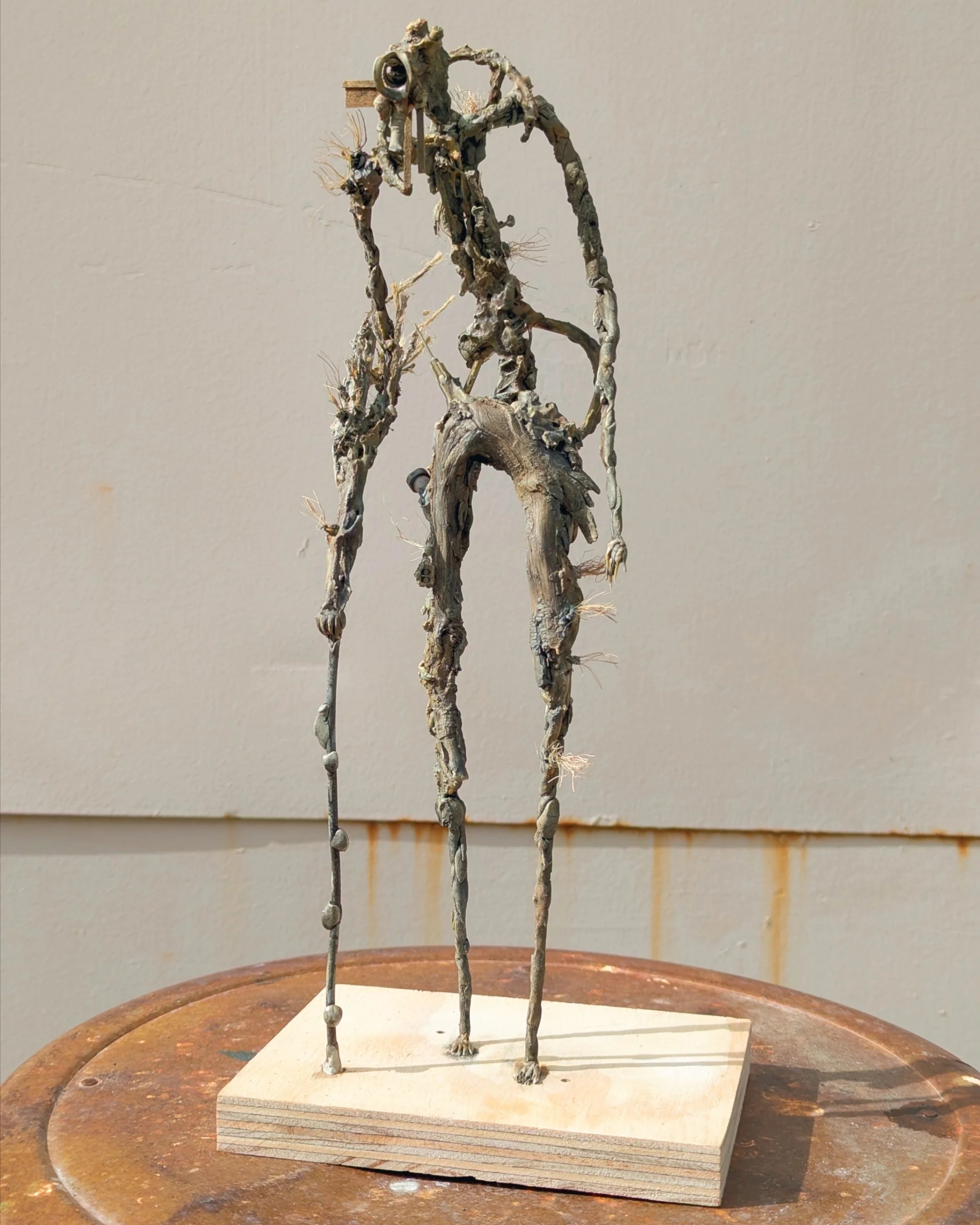 A sculpture made of metal wires and scrap pieces resembles a tall, abstract humanoid figure standing on a wooden base.