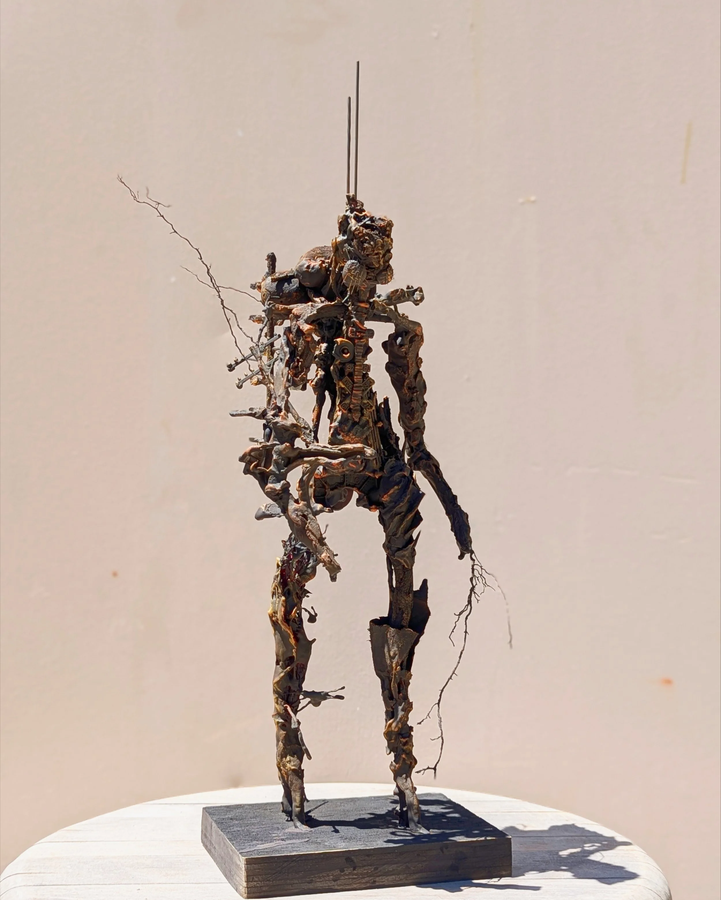 A sculpture resembling a humanoid figure with elongated limbs and a complex, textured surface, standing on a rectangular base on a white table.