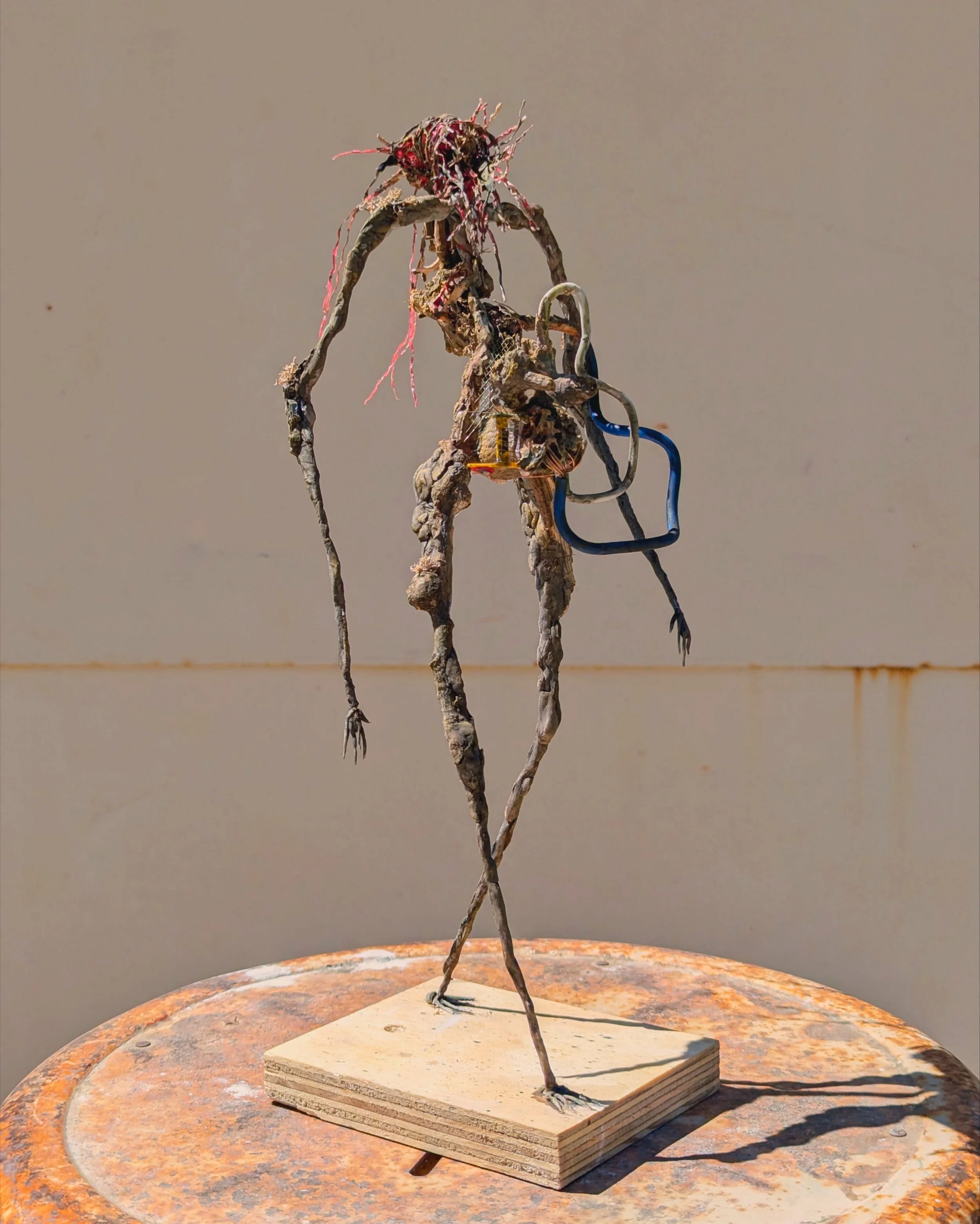 A sculptural artwork resembling a tall, thin humanoid figure made of twisted, rough materials. The figure has a head with pinkish, root-like hair, slender limbs, and is holding a small object. It stands on a wooden base on a rusty metal surface.