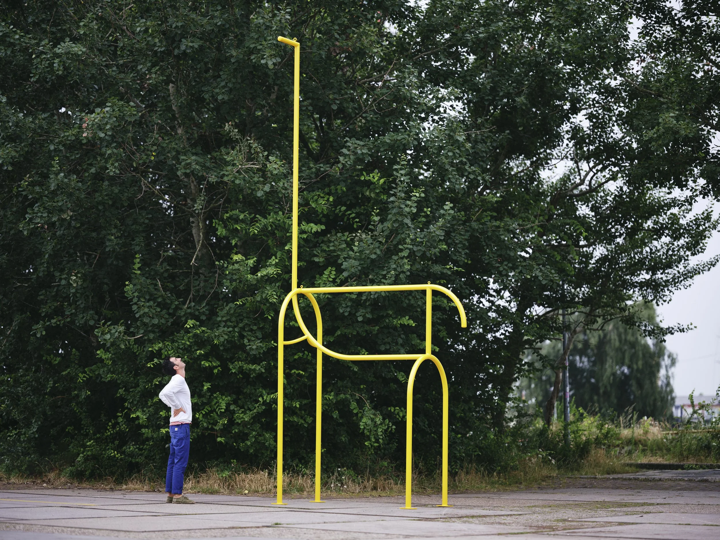 A man stands outdoors looking up at a yellow basketball hoop frame with no net, set in front of dense green trees on a cloudy day.