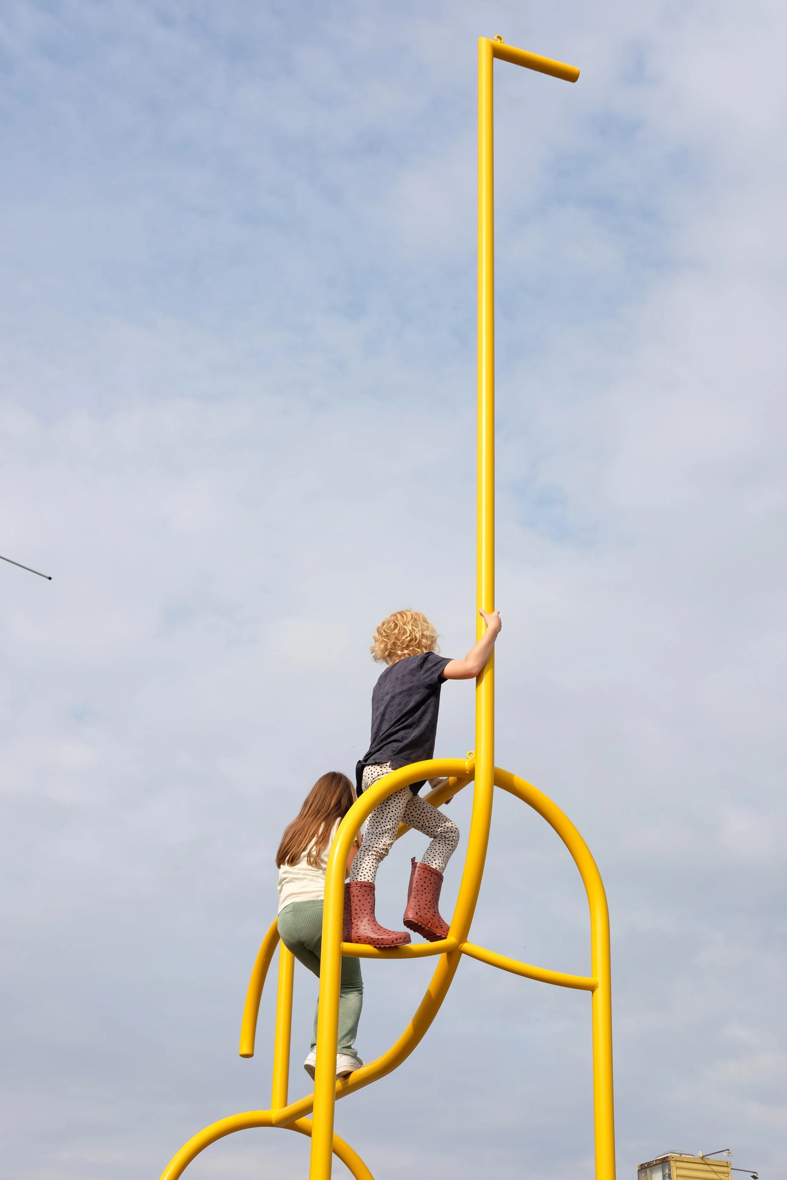 Two children climbing on a bright yellow playground structure with a pole, with a cloudy sky in the background.