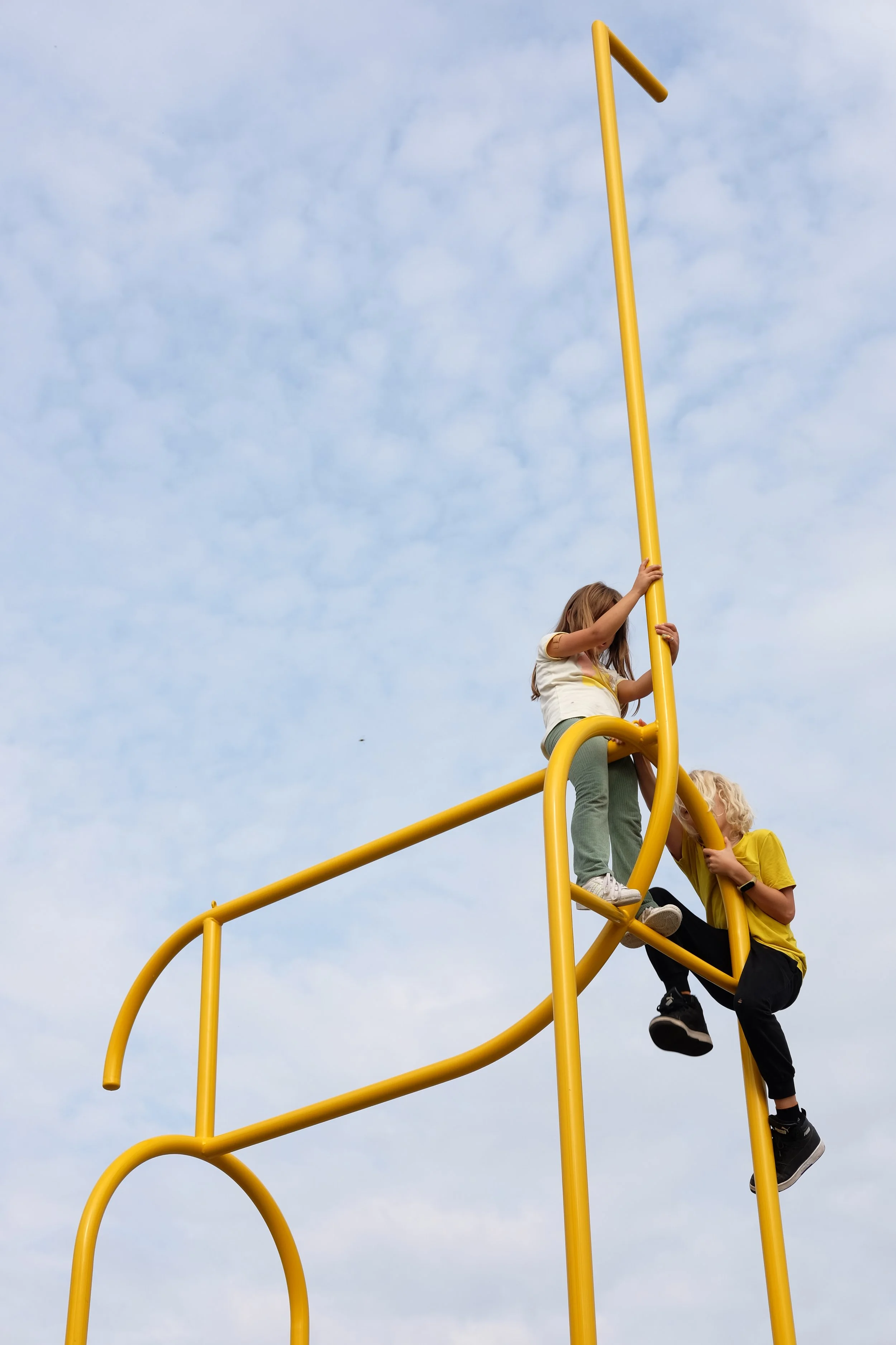 Two children playing on a yellow outdoor climbing structure against a cloudy sky.