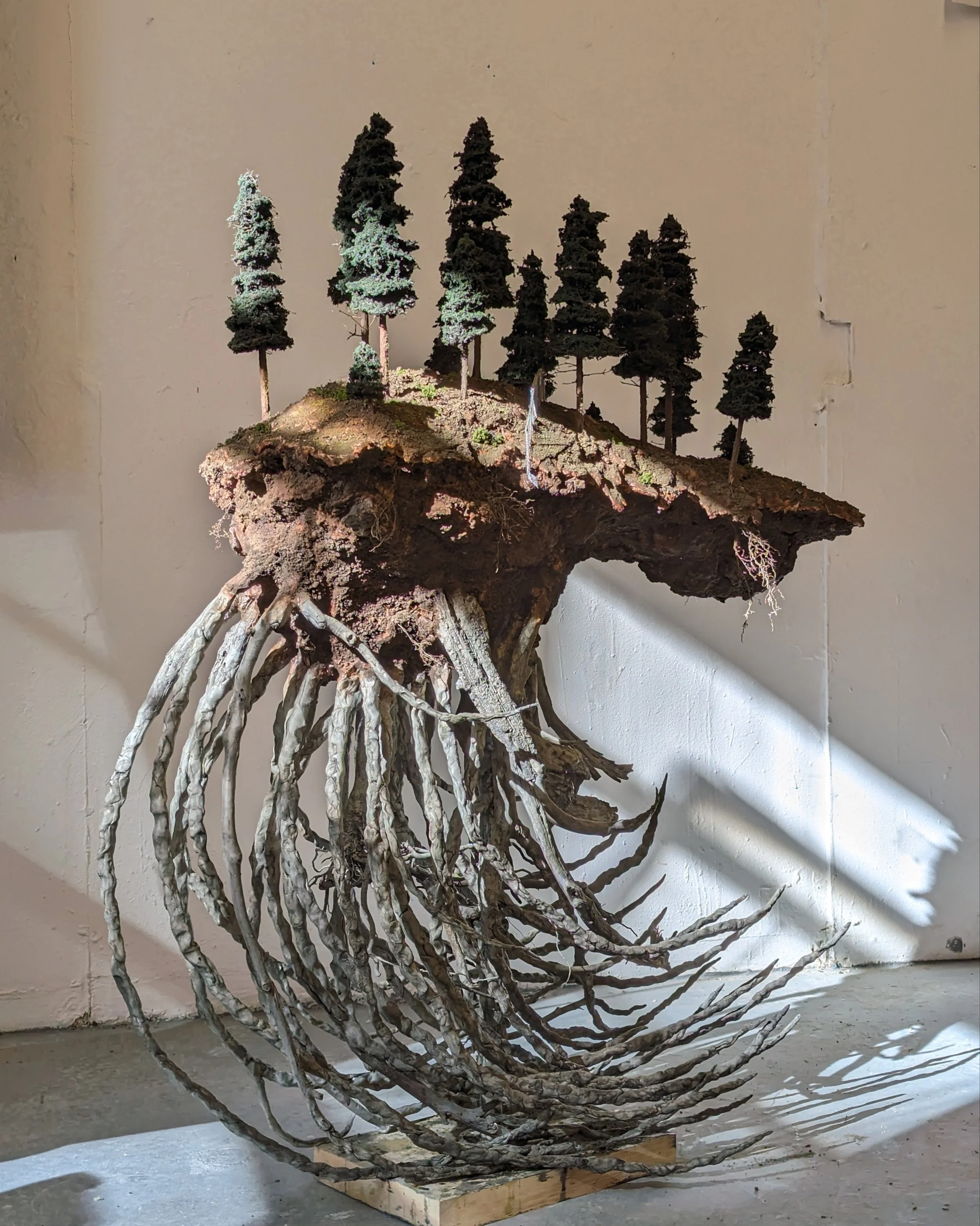 A large wooden sculpture resembling an upside-down mountain with trees on top, roots hanging down, placed on a wooden base, with shadows cast on the wall.