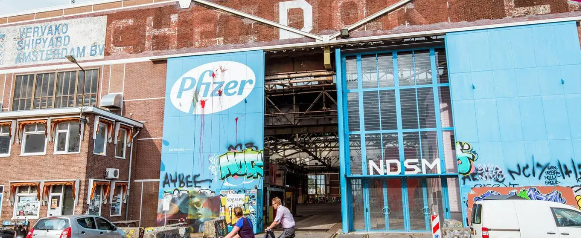 Street view of a large brick building covered in graffiti, with a large Pepsi logo on a blue banner and ND SM letters, with cars and pedestrians in front.