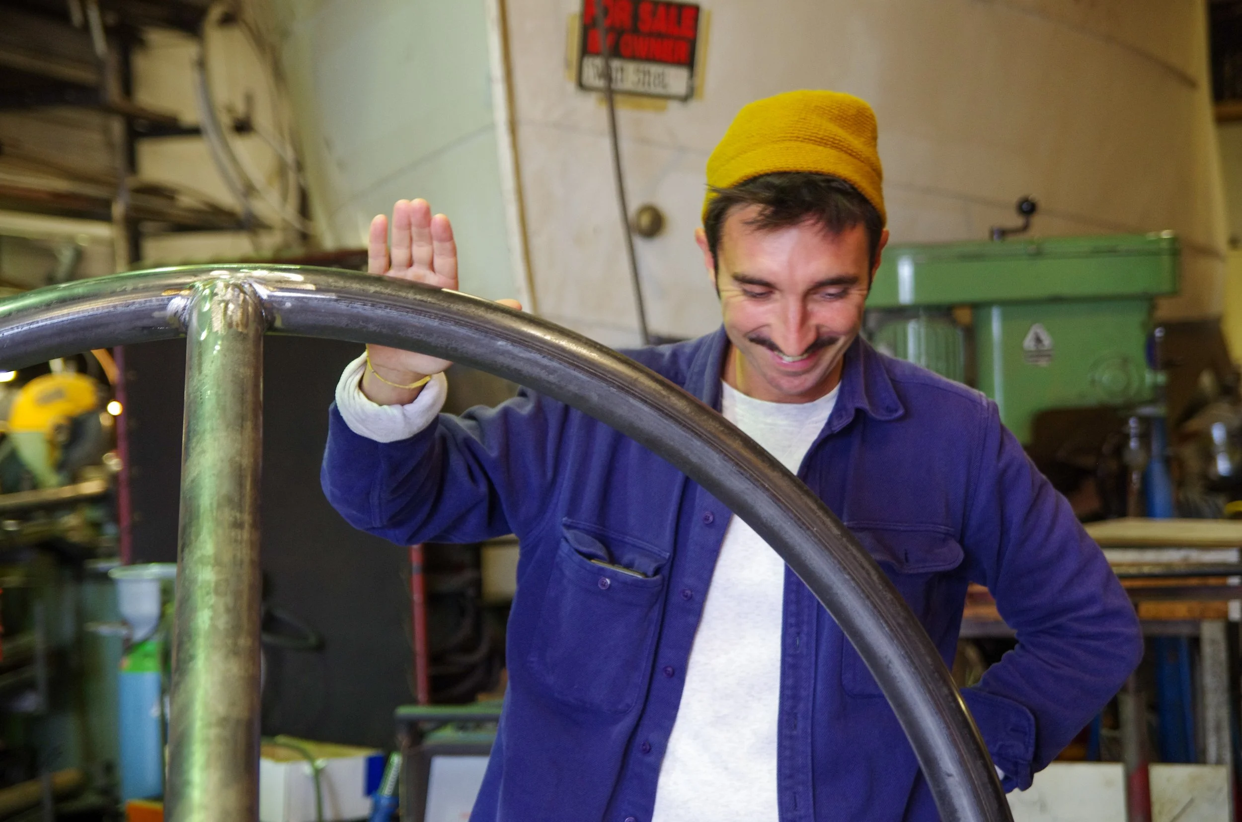 A man wearing a yellow beanie and a blue jacket, smiling, in a workshop, waving his hand.