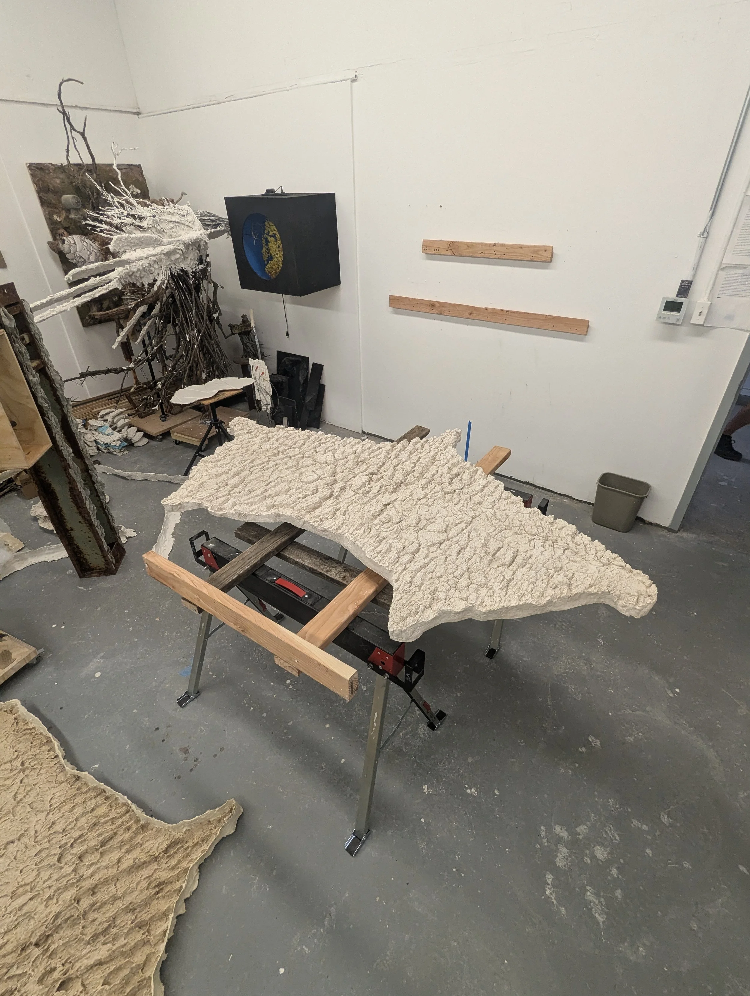 A workshop with a large piece of textured stone or ceramic on a portable workbench, surrounded by woodworking and sculpture materials, with wooden shelves on the white wall and a black cube artwork.