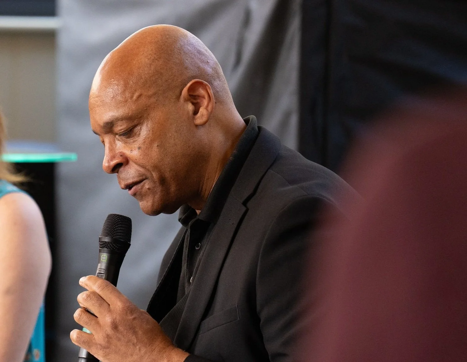 A man with a shaved head wearing a black suit holding a microphone and speaking or praying with eyes closed.