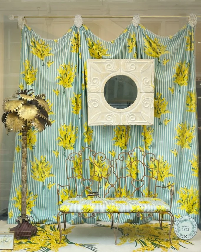 Décoration d'intérieur avec un rideau à rayures bleues et blanches avec des motifs floraux jaunes, un miroir carré blanc suspendu, un fauteuil en métal aux lignes courbes avec un coussin à motifs floraux, un lampadaire avec des feuilles dorées, et un tapis assorti aux motifs floraux jaunes.