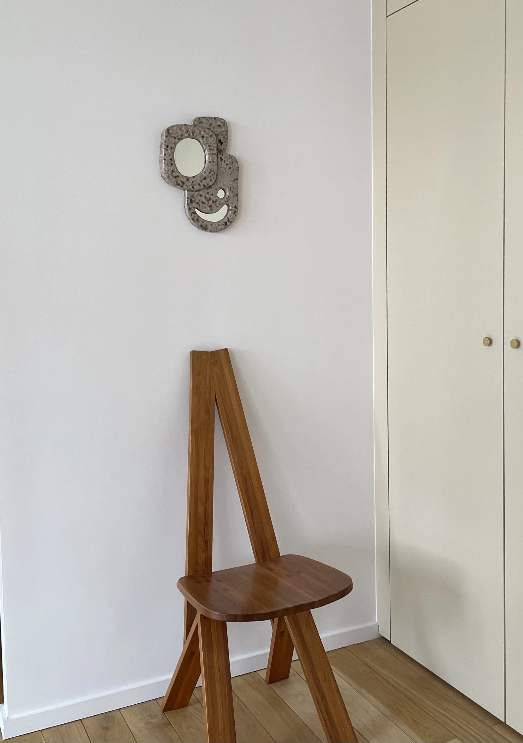 Projet de décoration d'intérieur pour un client privé. Une chaise en bois avec un mur blanc, décorée d'une sculpture murale en forme de visage stylisé modelé en céramique émaillée et d’une armoire blanche.