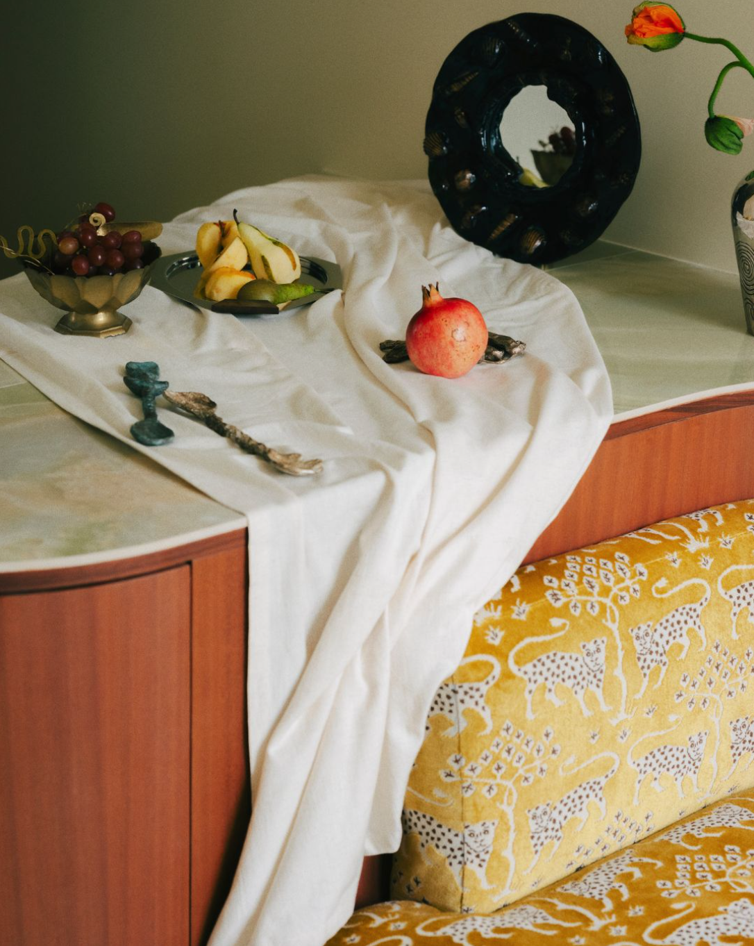 Une table de décoration avec des fruits, un miroir brun sombre en céramique avec des ornements marins fait de corde et de coquillage, une pomegranate, et un vase avec une fleur, sur une nappe blanche. Projet architectural de Batik Studio.