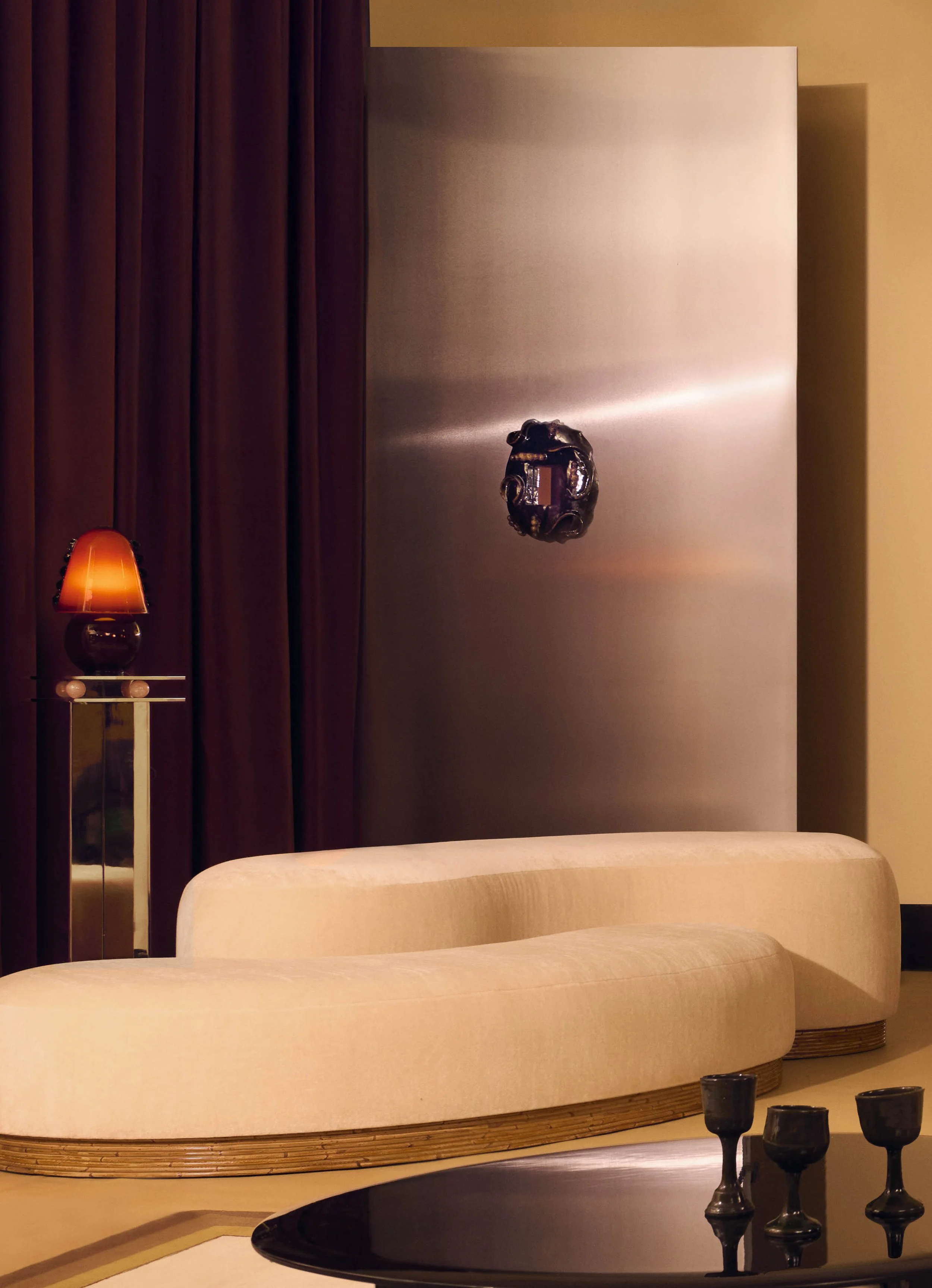 Un salon avec un canapé beige arrondi, une table basse noire avec des verres, un mur en métal avec une plaque décorative, et un lampadaire avec une lampe orange à gauche. Publié dans le magazine Elle décoration, article par Aurore Sfez.