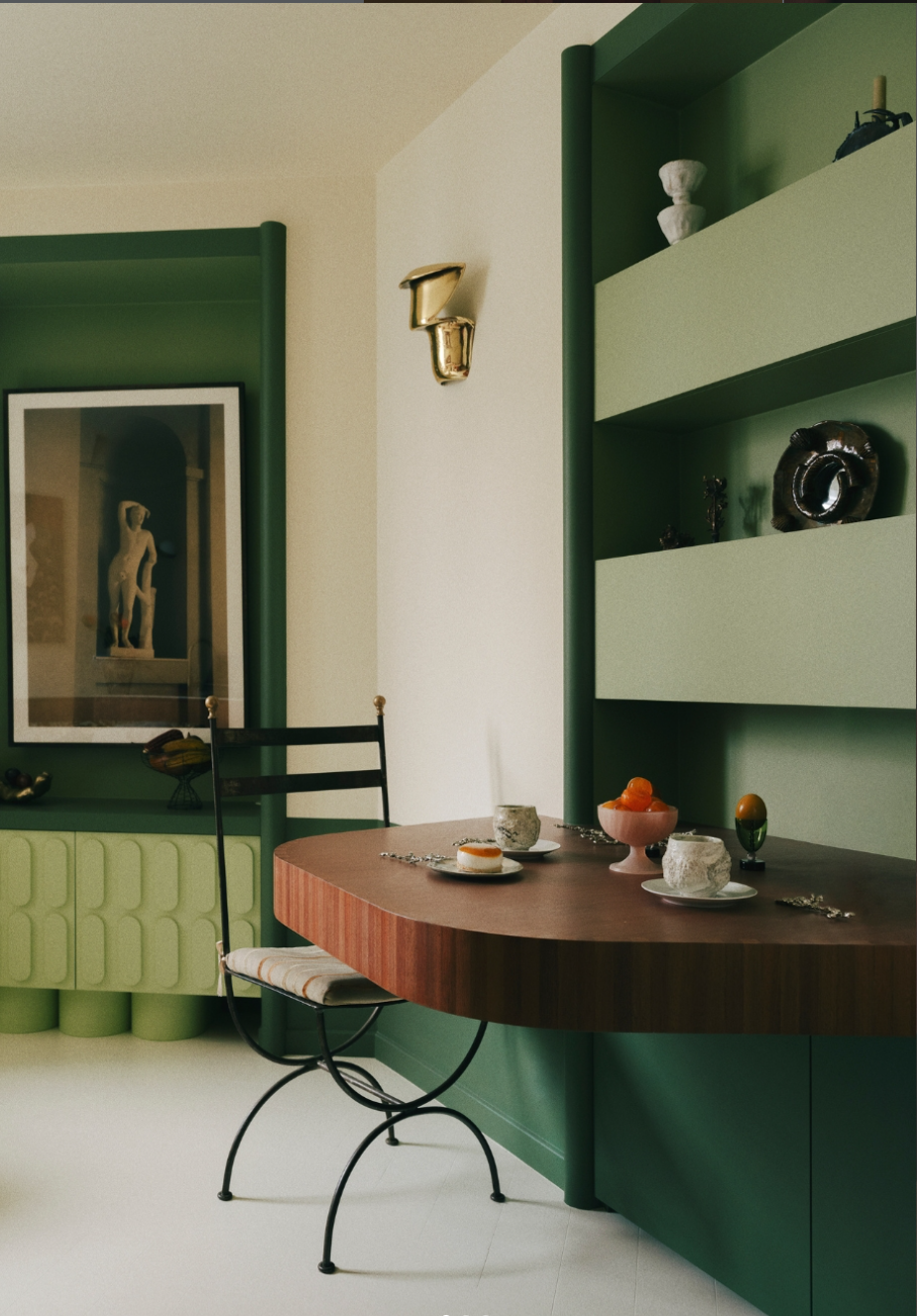 Intérieur d'une salle à manger avec une table en bois, décorée de vaisselle et d'objets décoratifs, murs verts avec étagères intégrées et une œuvre d'art visible dans un cadre. Dont un miroir brun avec des ornements de coquillages. Archi Batik Studio