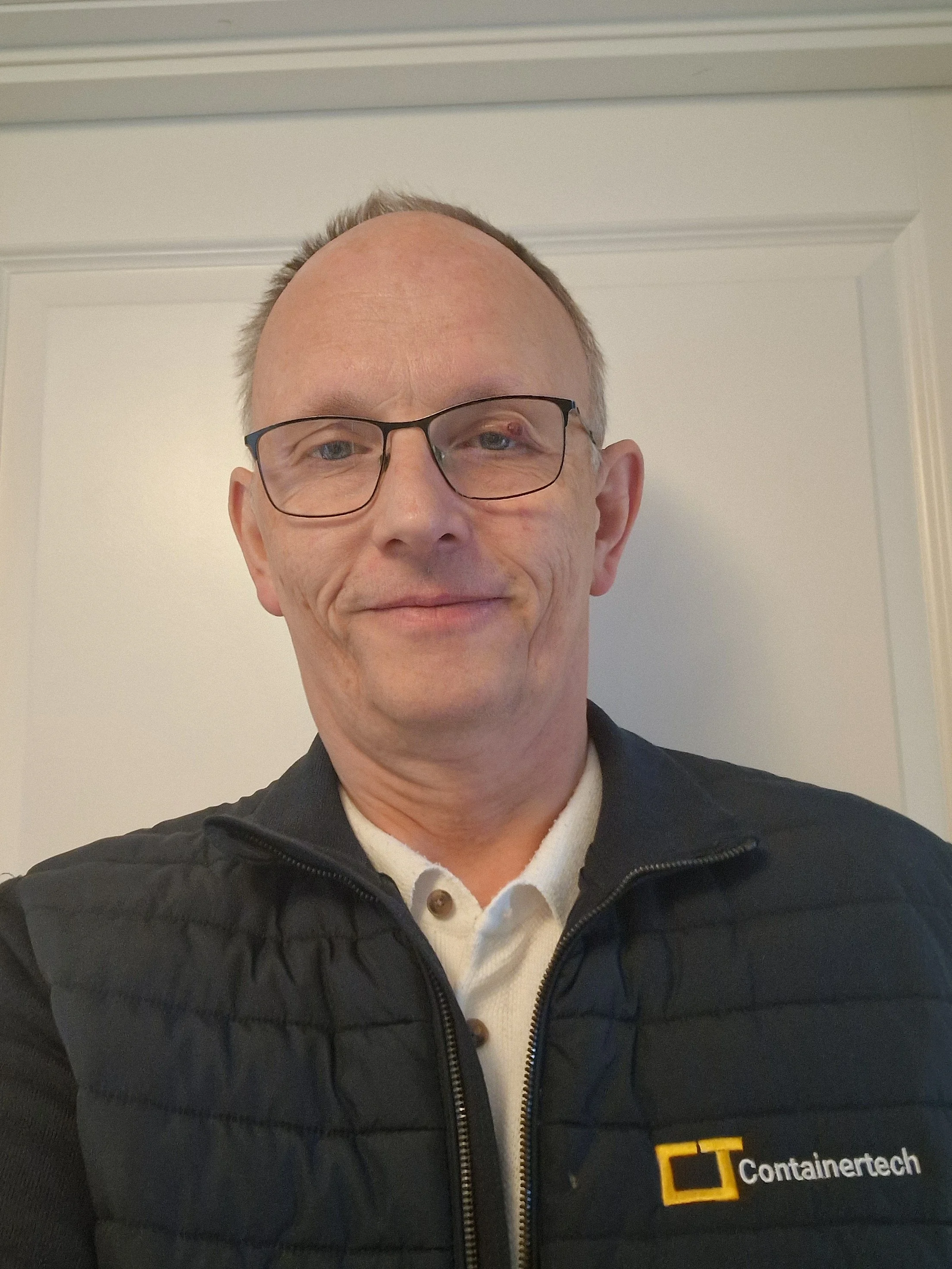 Bilde av en mann med briller, kort hår, smilende, iført hvit skjorte, mot en beige vegg.