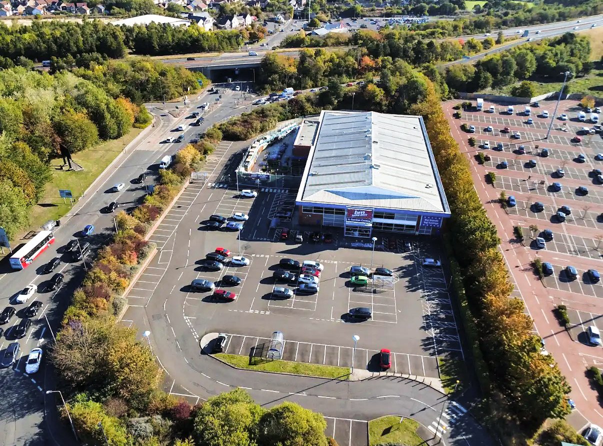 Retail warehouse with a large black Mercedes logo sign, building, cars, vans, and trucks outside, and cloudy sky.