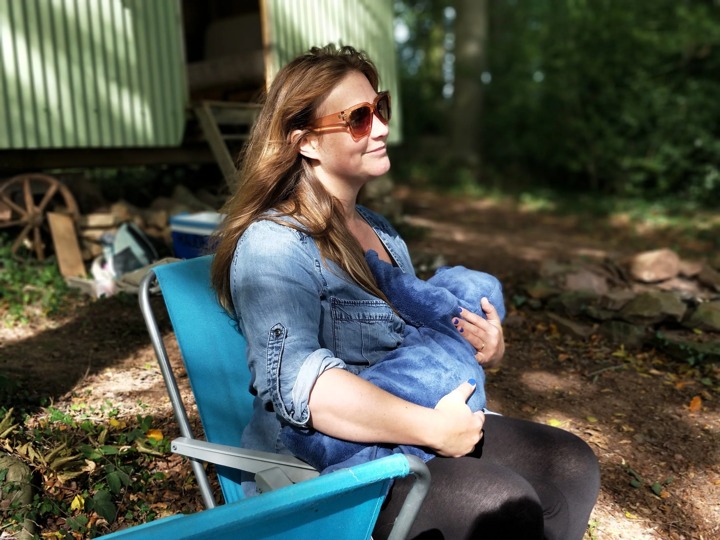 Woman sitting in a chair outdoors holding a baby wrapped in a blue blanket, wearing sunglasses and a denim shirt.