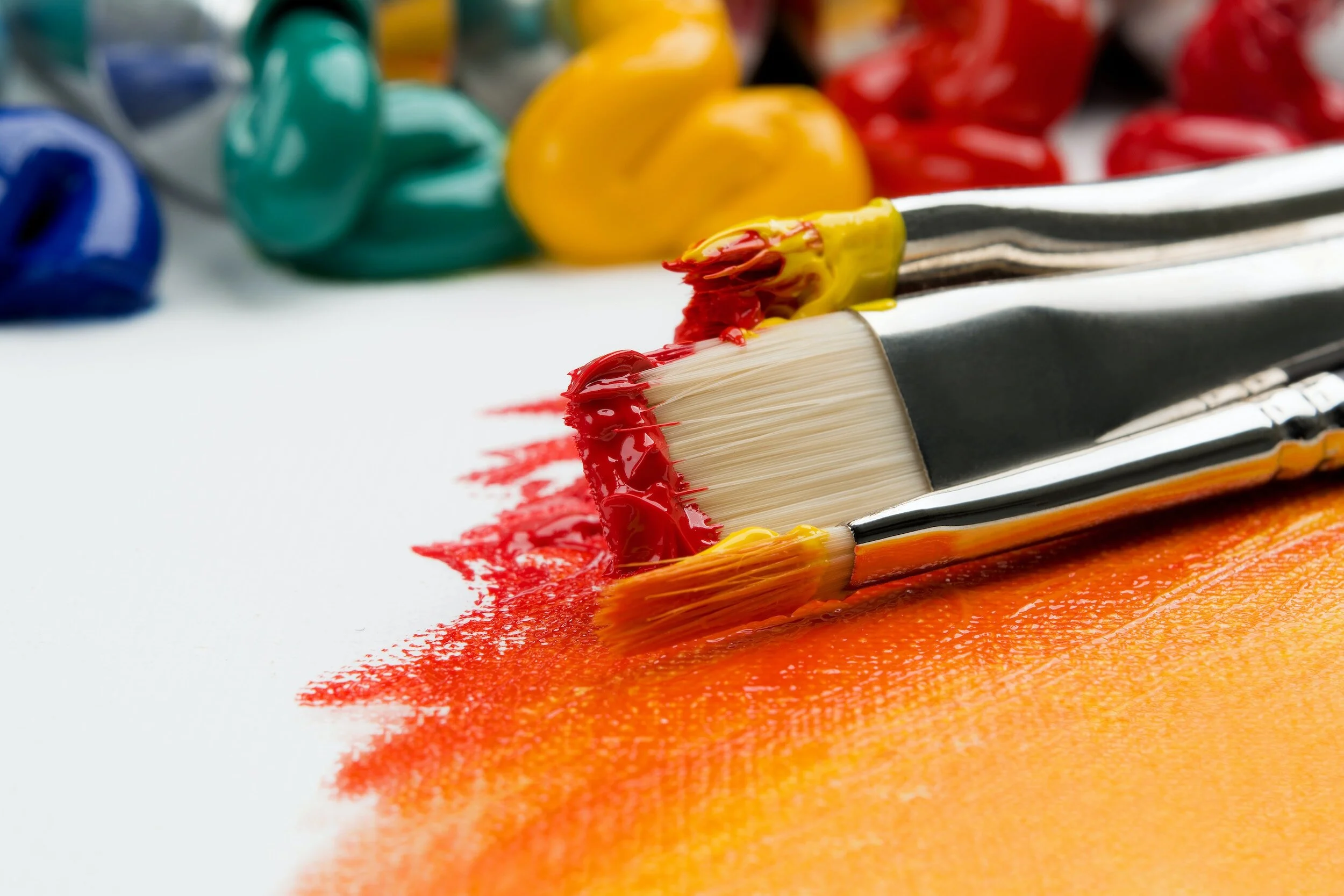 Paintbrushes with red and yellow paint on canvas, surrounded by tubes of colorful paint.