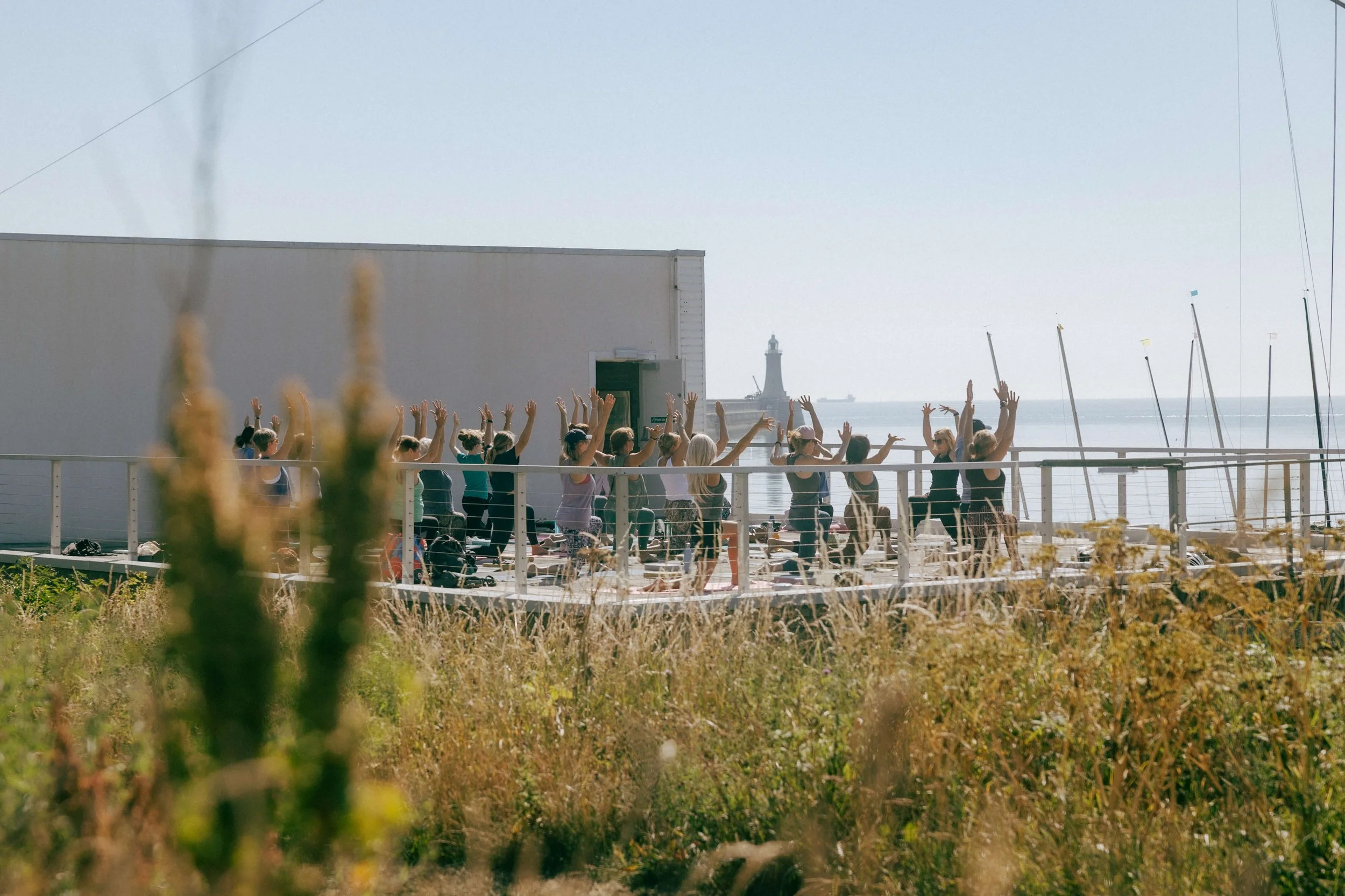 Group of people practicing yoga outdoors near the water with a lighthouse in the background at Jasmine Yoga Tynemouth