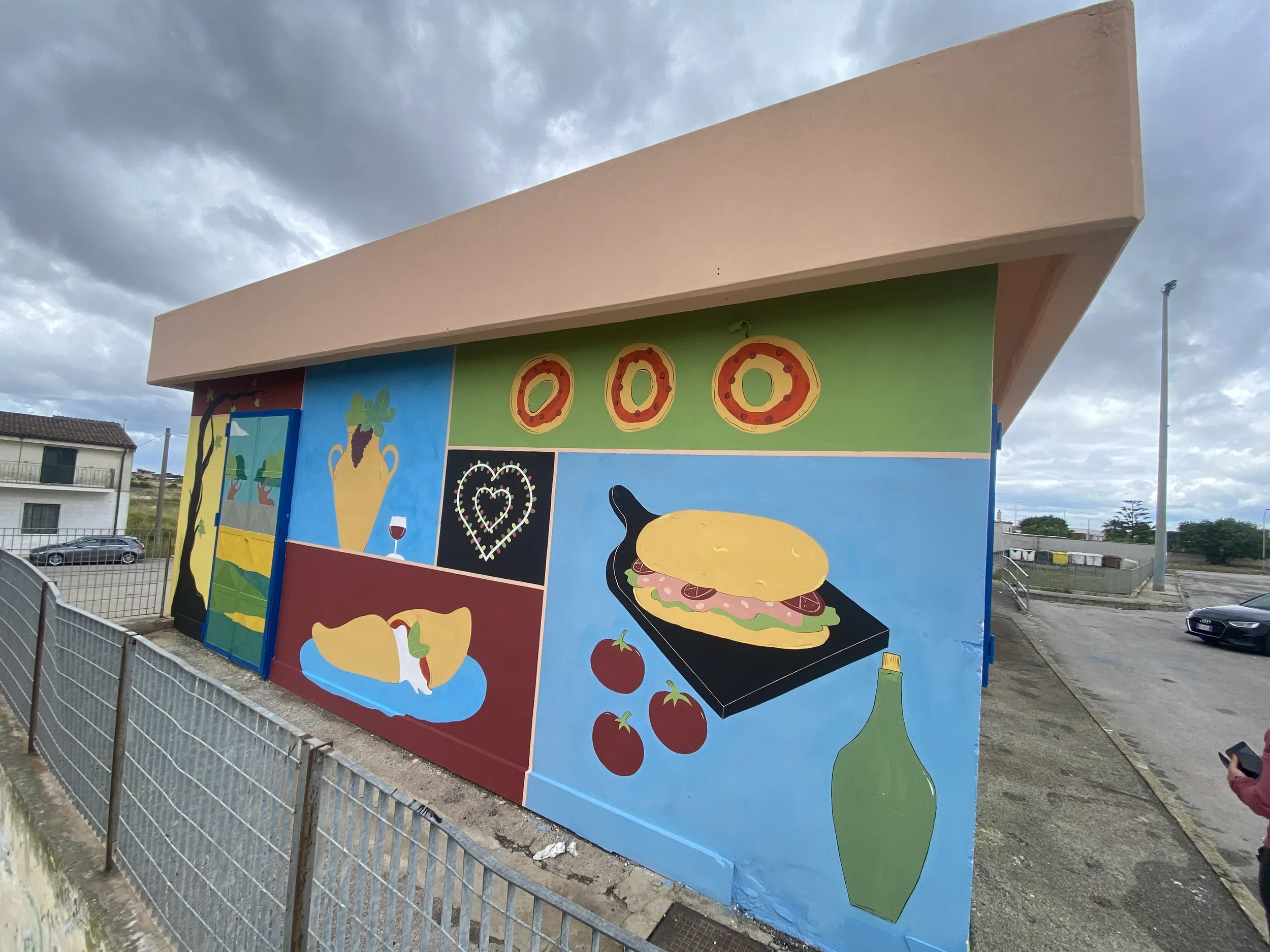 Colorful mural on an outside building depicting various food items including pizza, a sandwich, tomatoes, a bottle, and a wine glass, with a sky and some houses in the background.