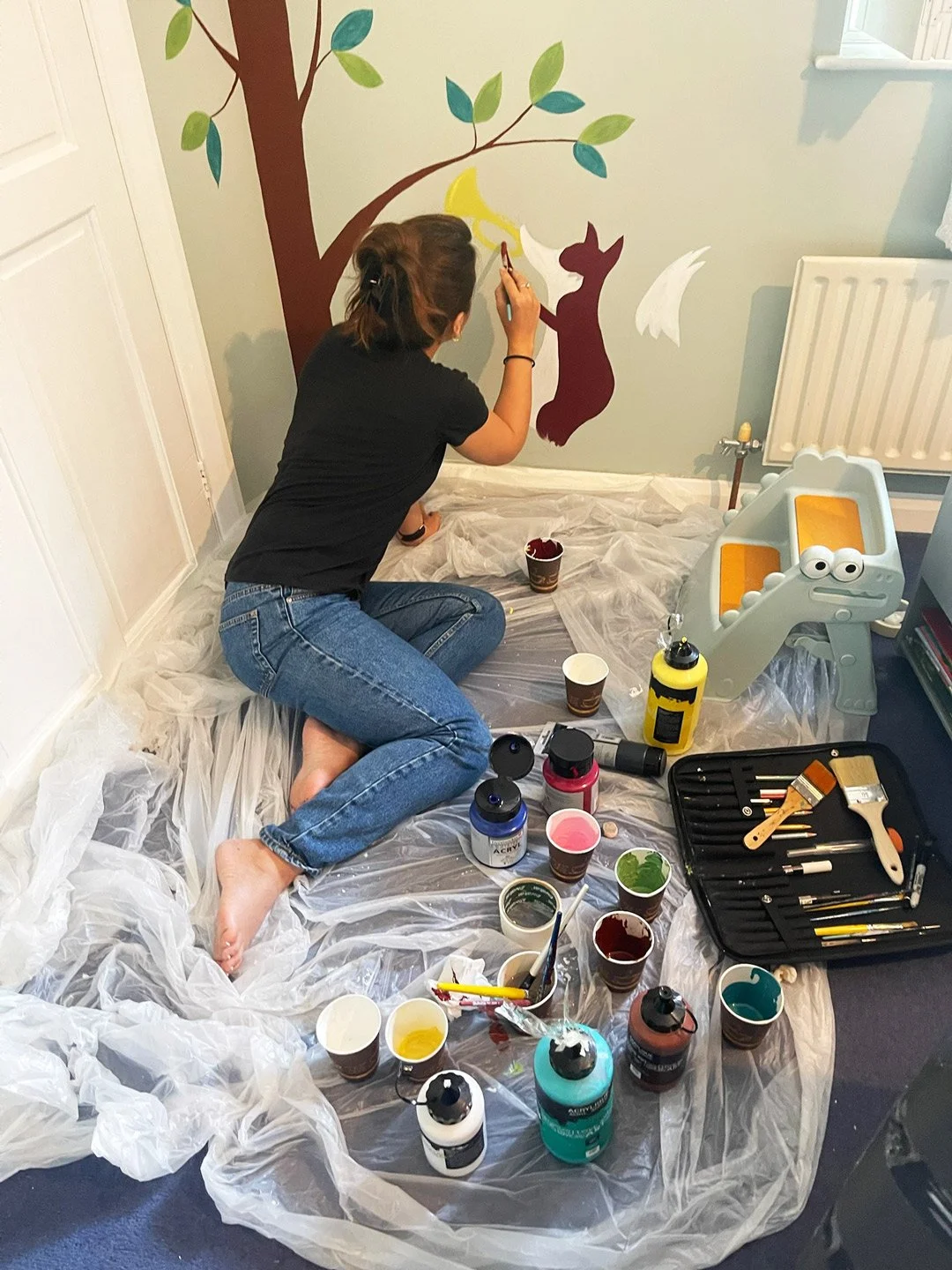 Woman painting a mural of a tree with leaves, a fox, and a moon on a light-colored wall, surrounded by painting supplies and plastic sheeting on the floor.