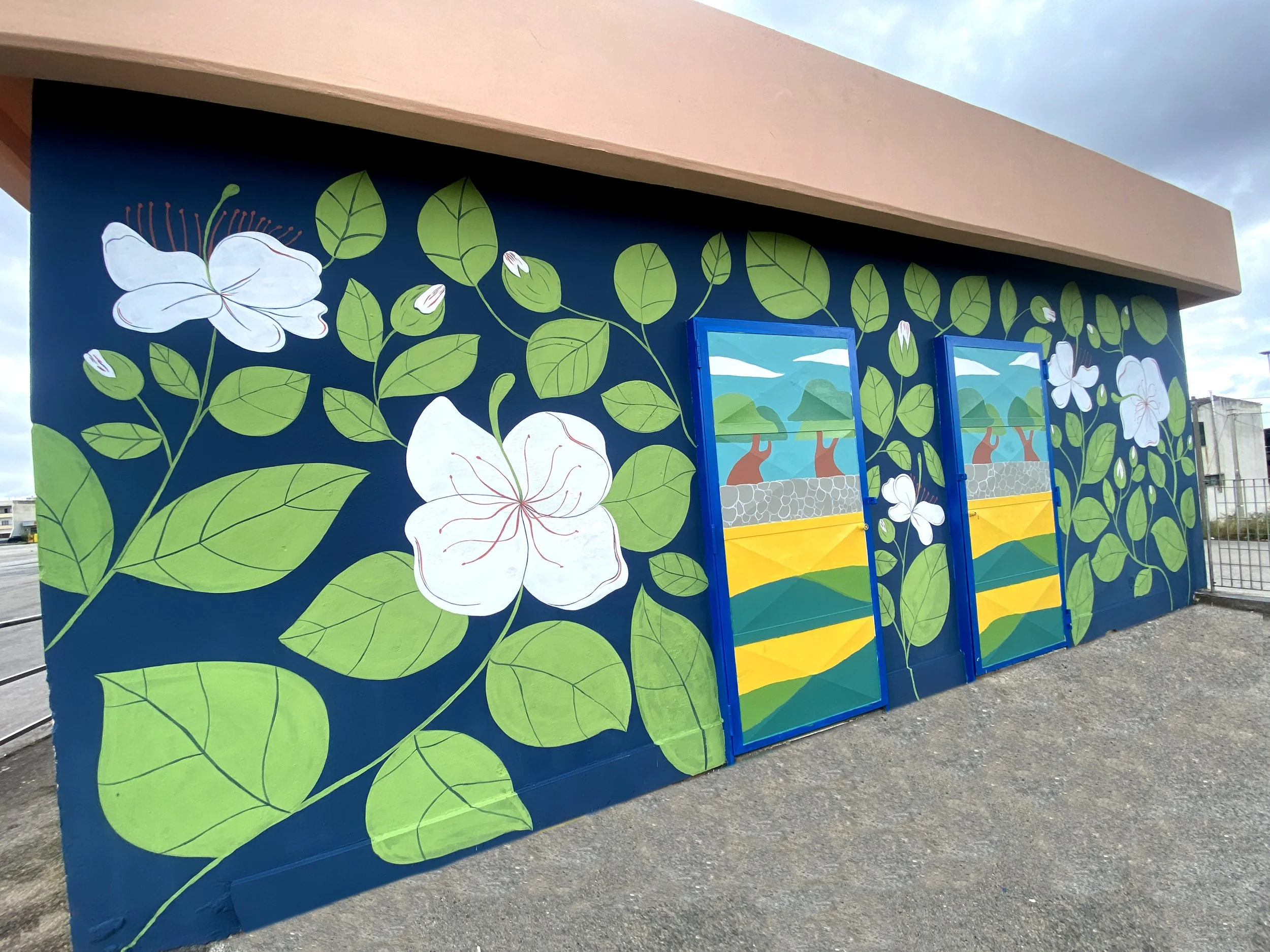 Wall mural with white flowers, green leaves, and two brightly painted doors depicting a landscape with mountains, water, and trees.