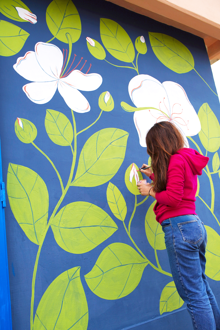 Woman painting white flower with pink and red details on a blue wall featuring large green leaves and flower buds.