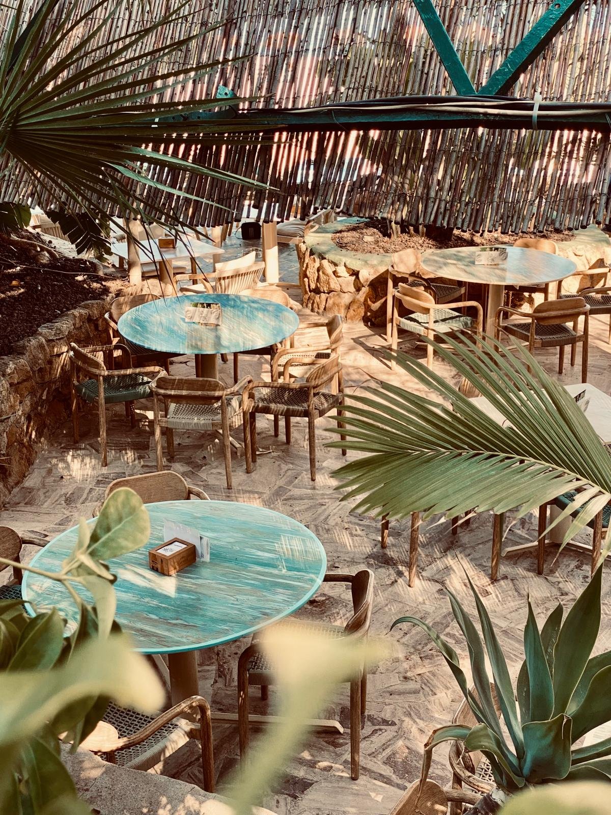 Terraza al aire libre con mesas redondas y sillas de mimbre, rodeada de plantas tropicales y cubierta de bambú.