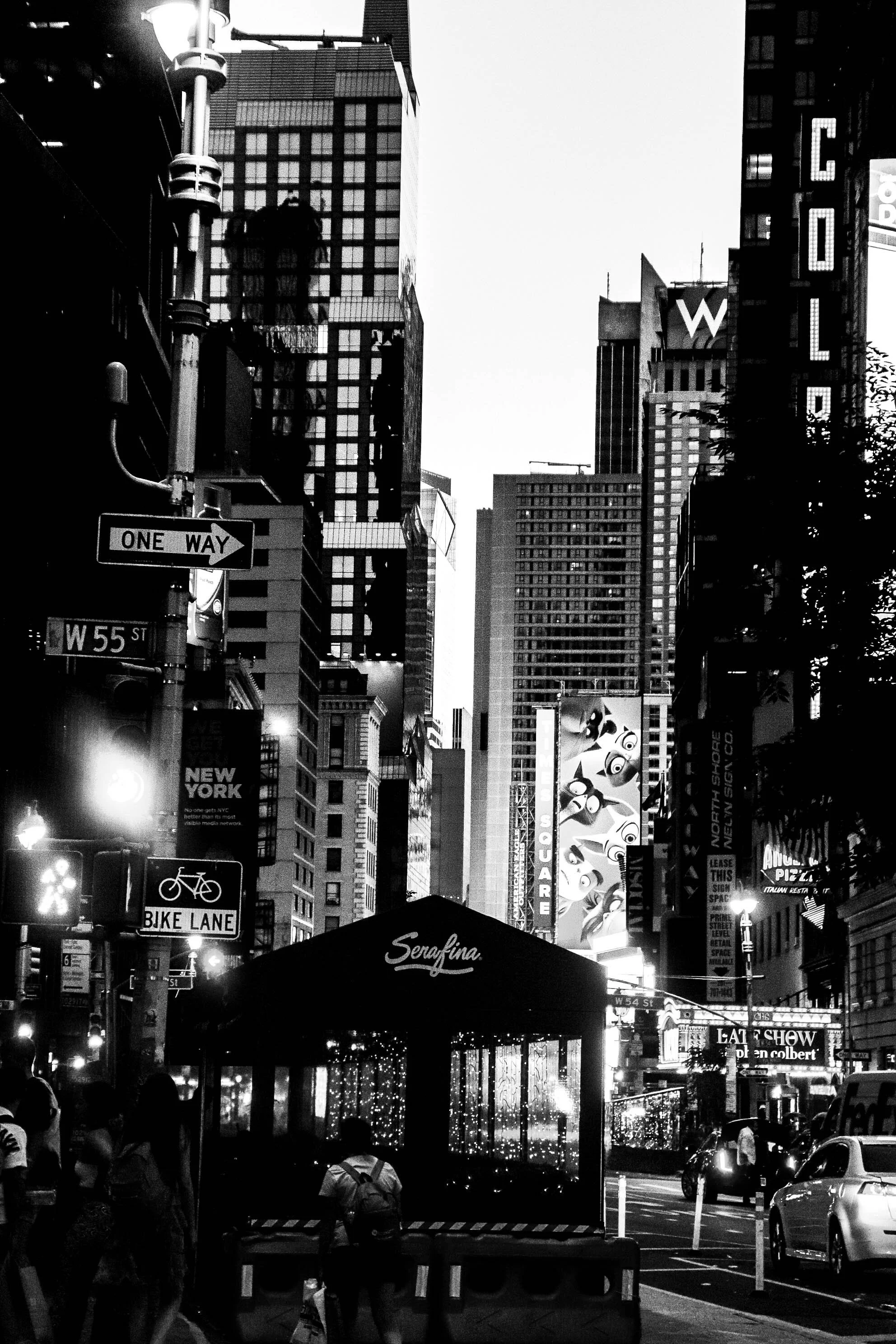 A black and white photo of Times Square in New York City showing tall buildings, billboards, traffic, pedestrians, and street signs including a bike lane and one-way street sign.