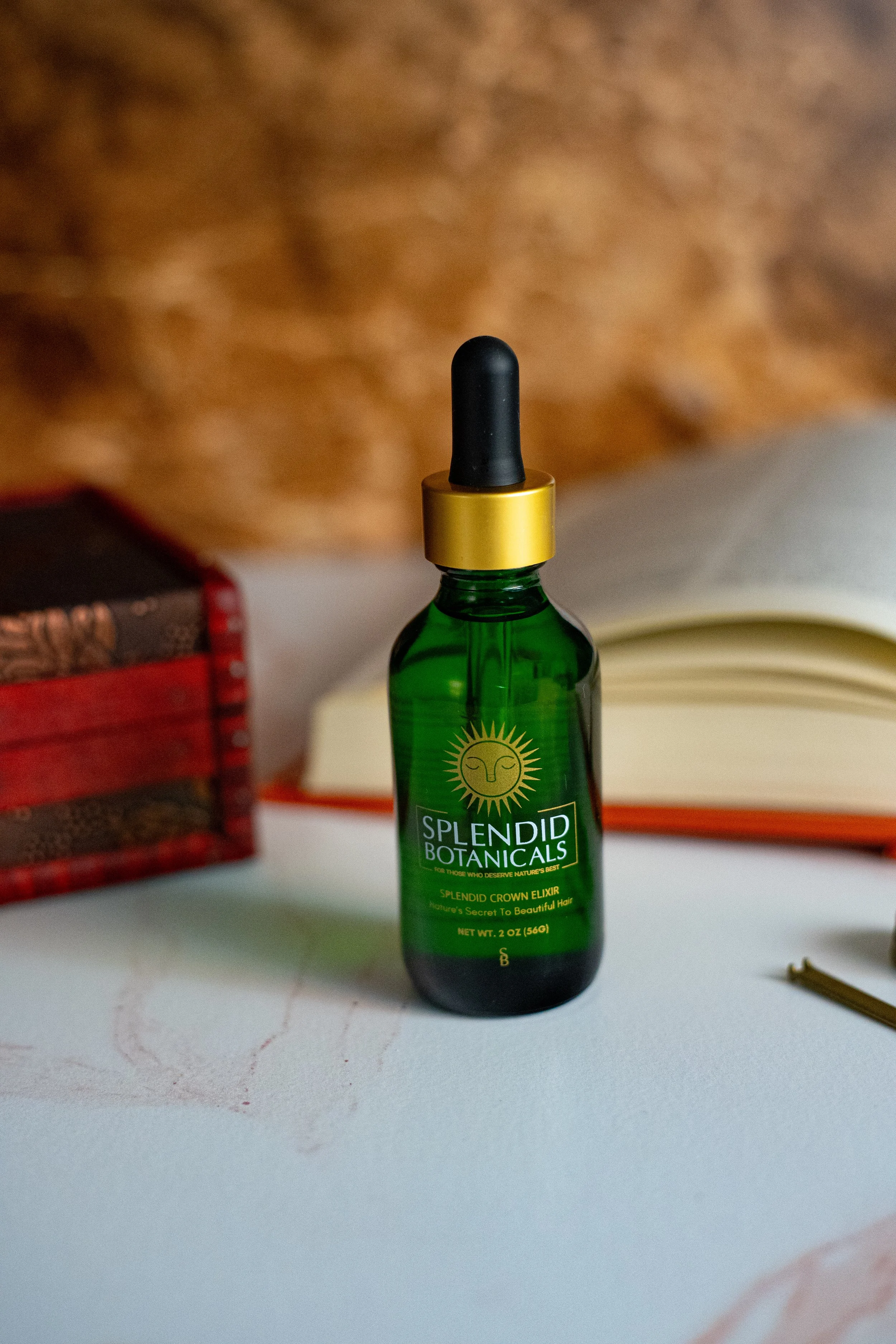 A green bottle of Splendid Botanicals Crown Elixir hair product with a black dropper cap on a white surface, with books in the background.