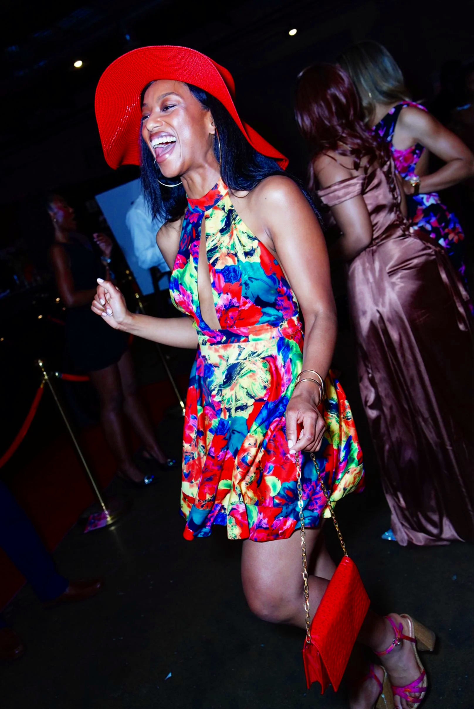 A woman wearing a colorful floral dress and a large red hat, dancing and laughing at a lively event with other women in the background.