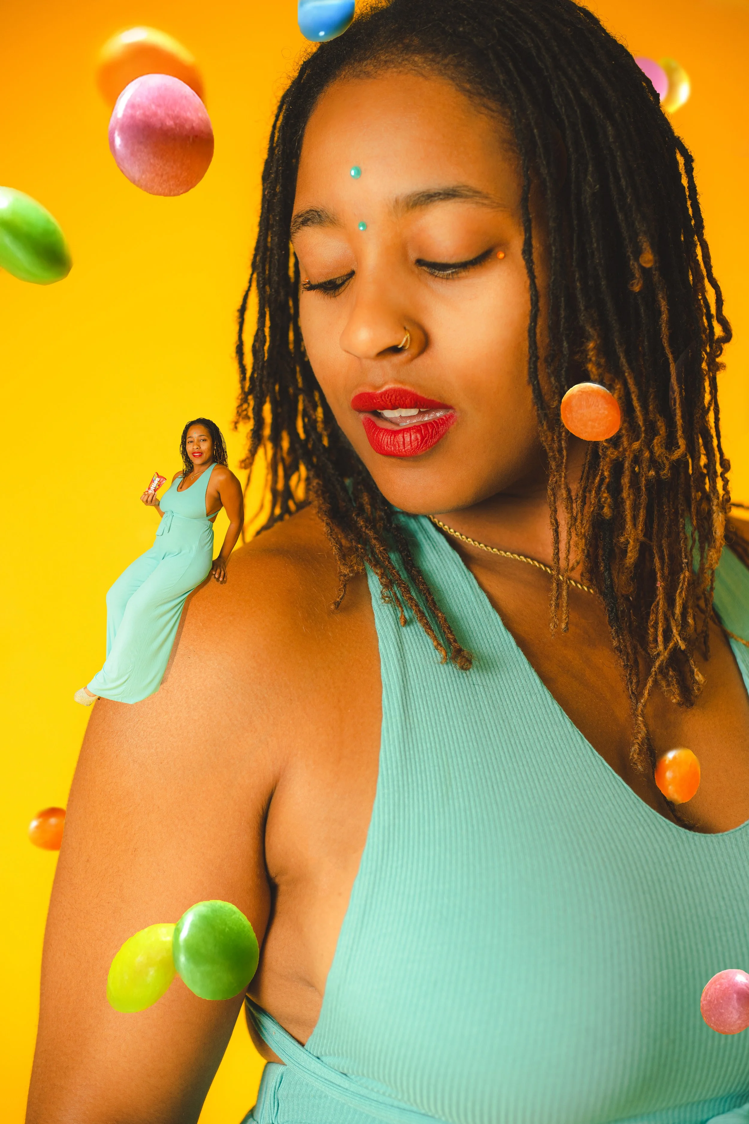 A woman with long dreadlocks wearing a teal sleeveless top, with colorful floating candies around her against a yellow background.