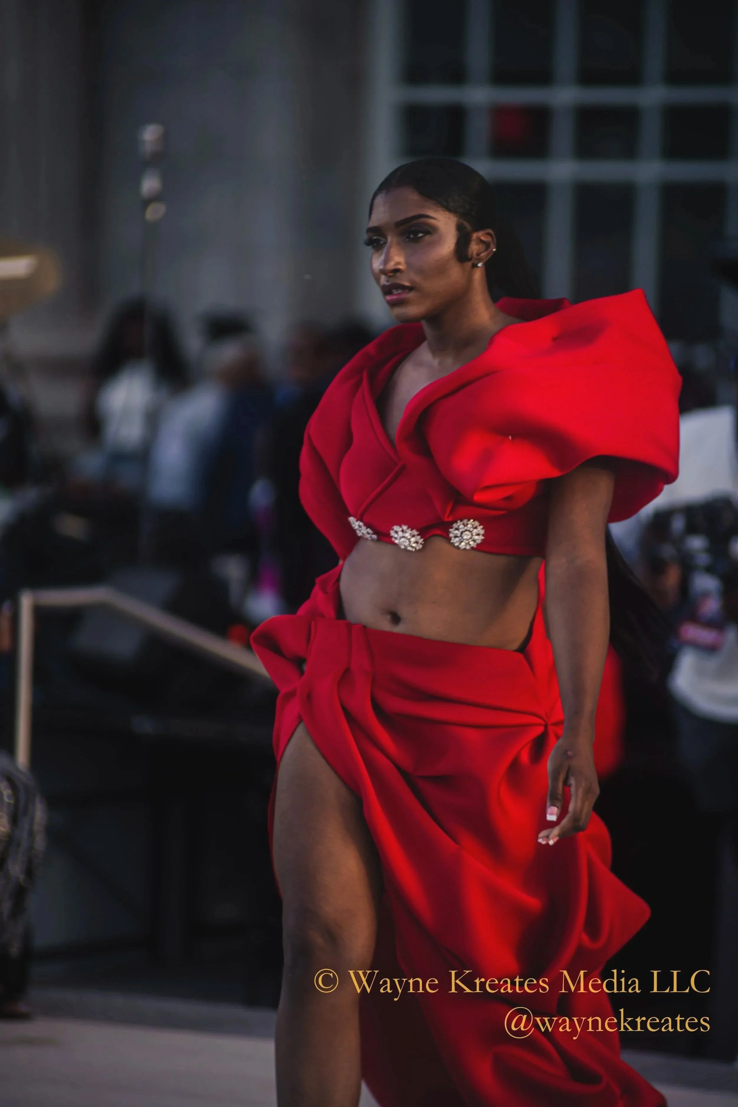 A woman wearing a striking red outfit with puffed sleeves and a skirt, strutting on a runway outdoors in an urban setting at dusk.