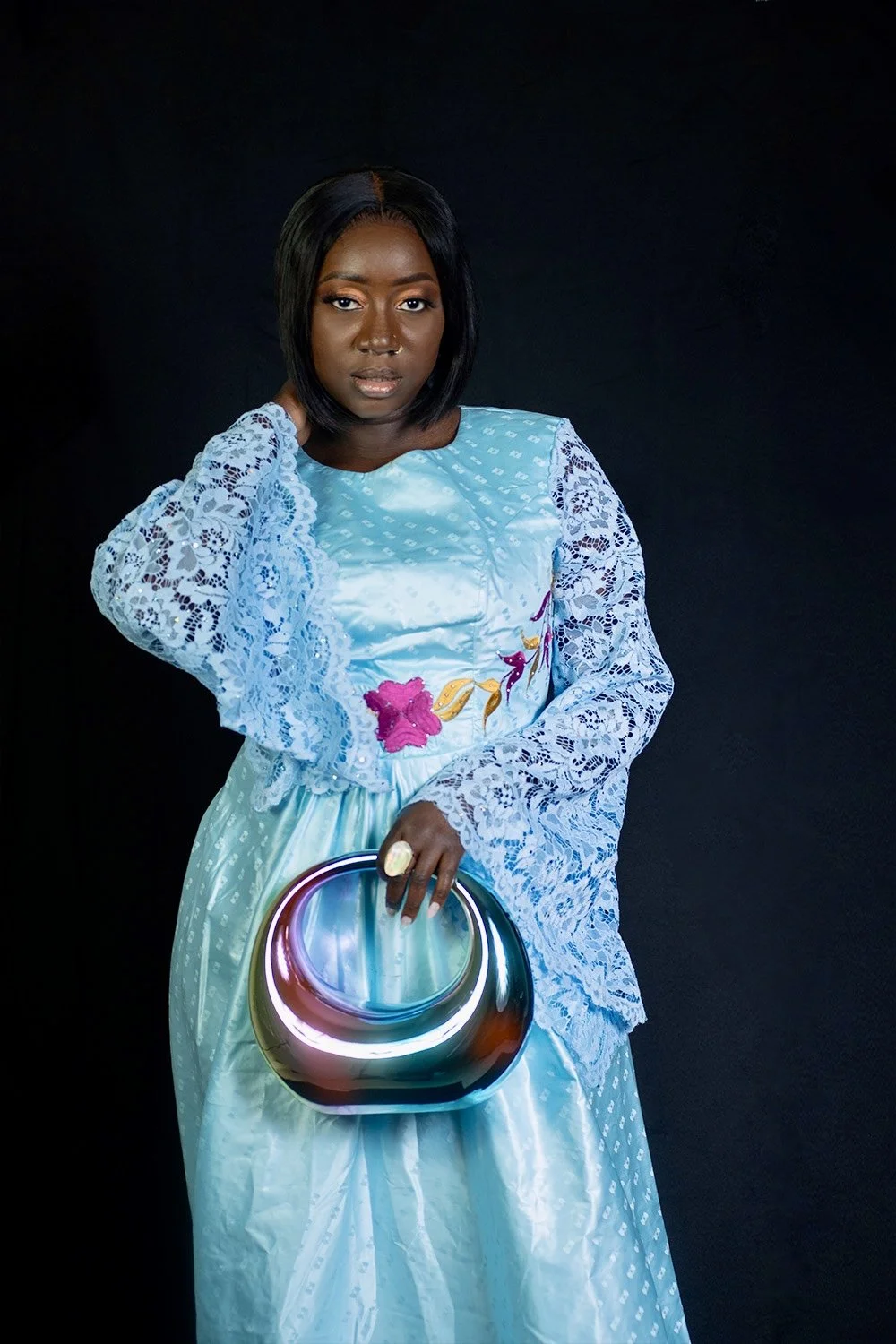 A woman with dark hair posing against a black background, wearing a blue dress with lace sleeves and floral embroidery, holding a reflective clutch purse.