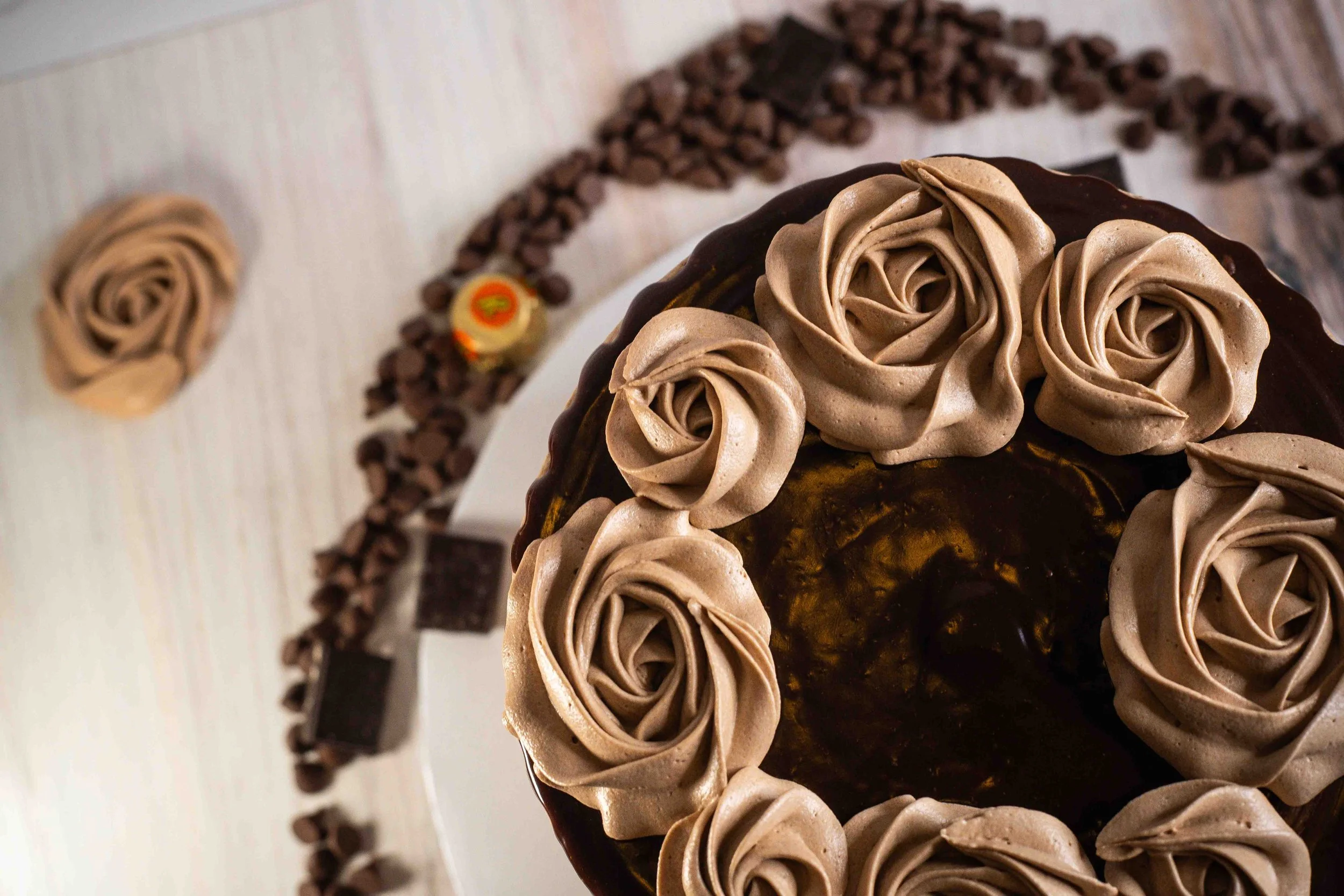 Close-up of a chocolate cake decorated with chocolate icing roses on top, surrounded by a ring of chocolate chunks, with scattered chocolate pieces on the table.