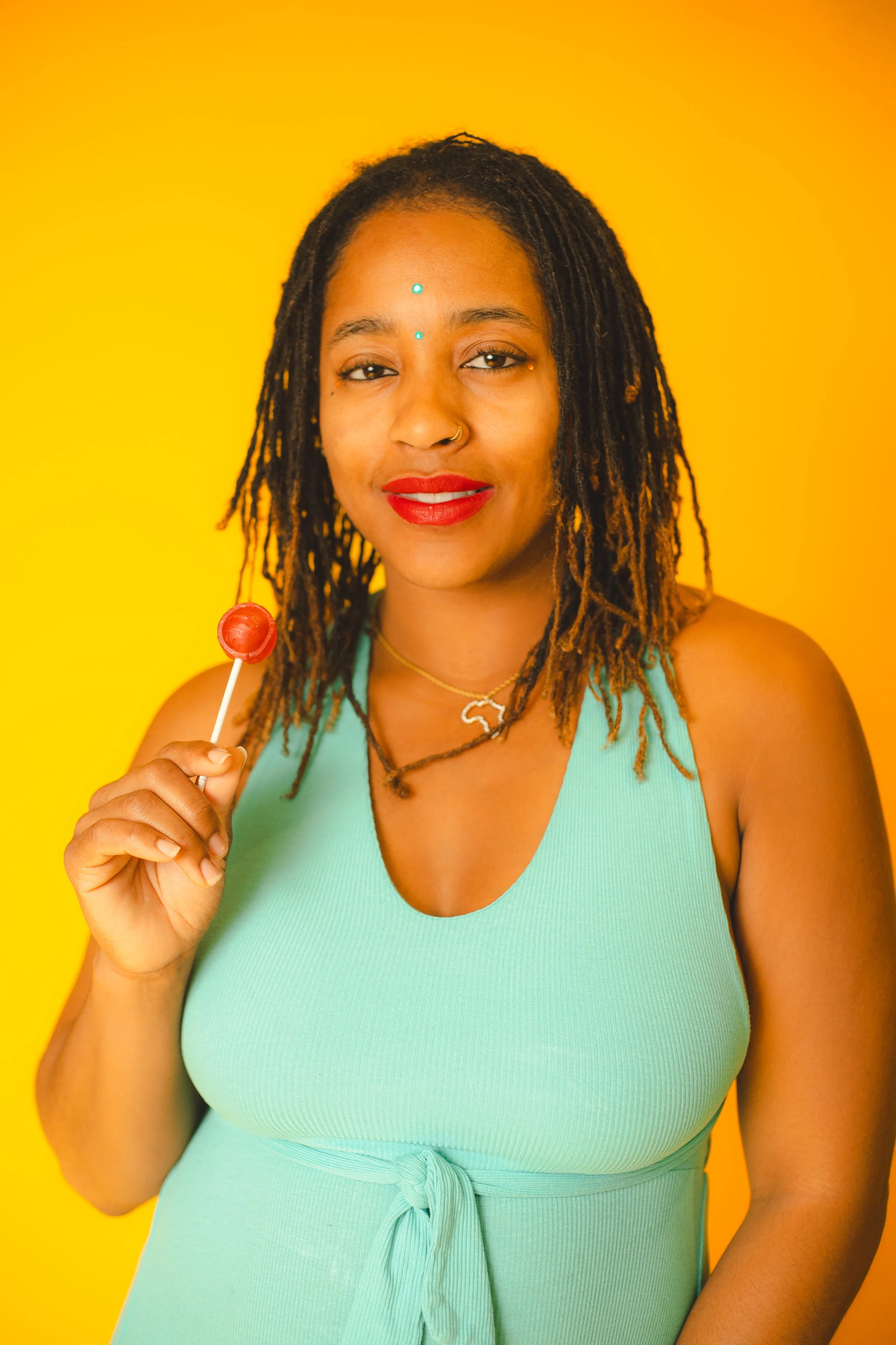 A woman with dreadlocks and red lipstick holding a red lollipop, wearing a mint green dress, and standing against a yellow background.