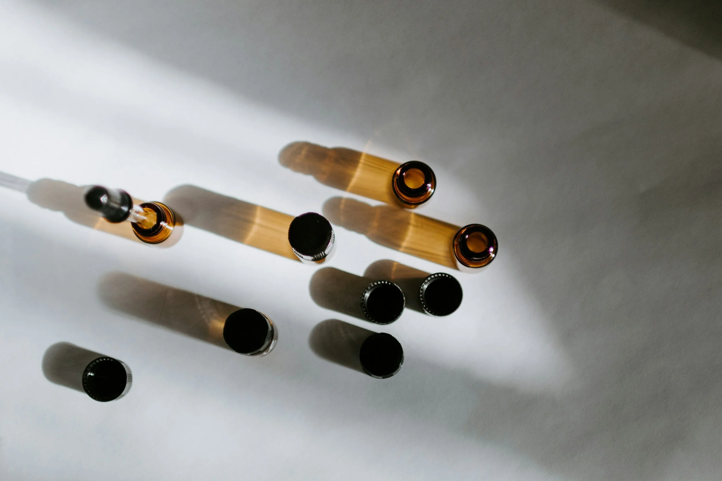 Top-down view of various small glass bottles with black caps, casting elongated shadows on a white surface, illuminated from the left side.