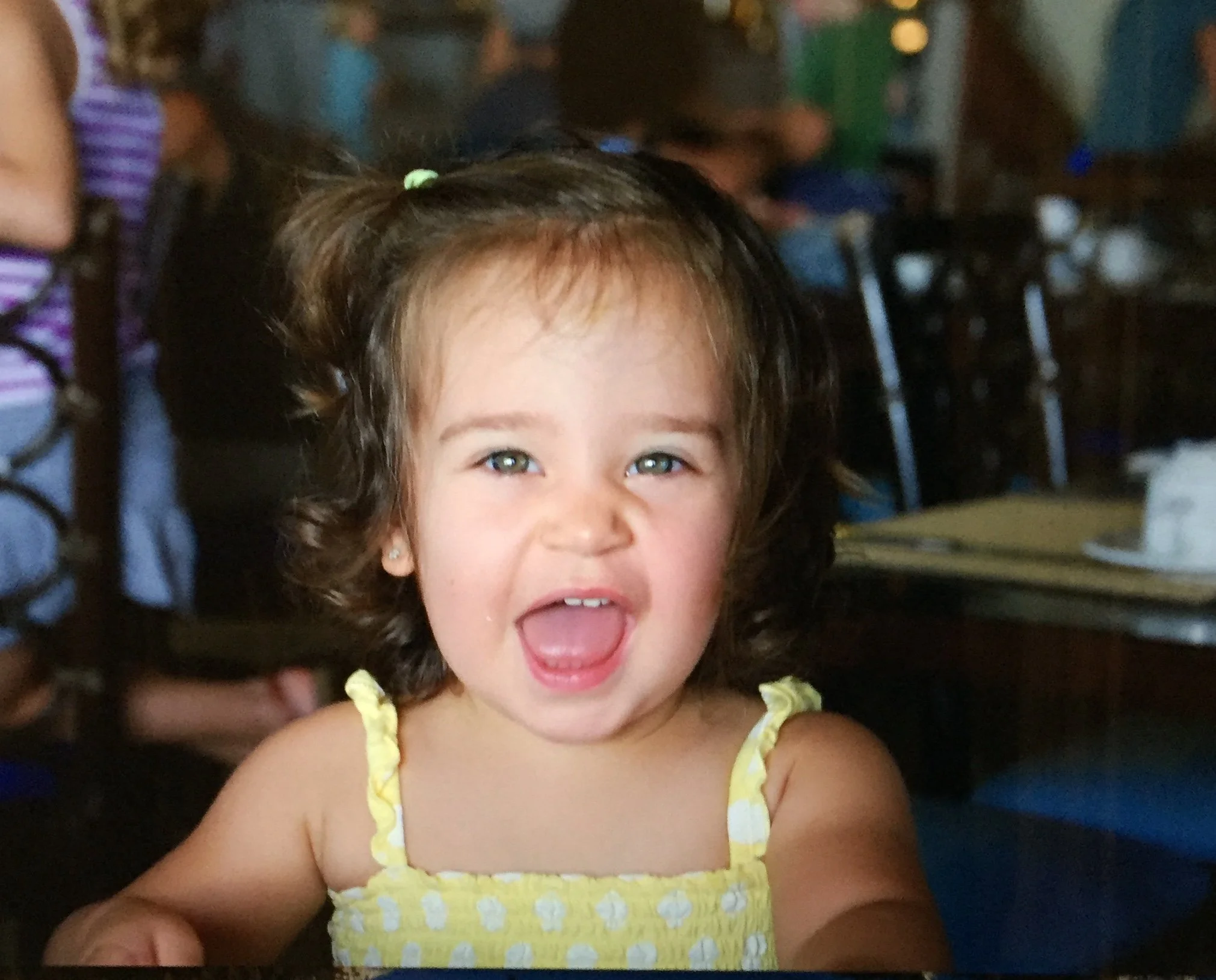 Young Andrea Coppé with brown curly hair, wearing a yellow dress with white polka dots, joyfully smiling with her tongue slightly sticking out. The background shows a room with many people, some sitting on chairs.