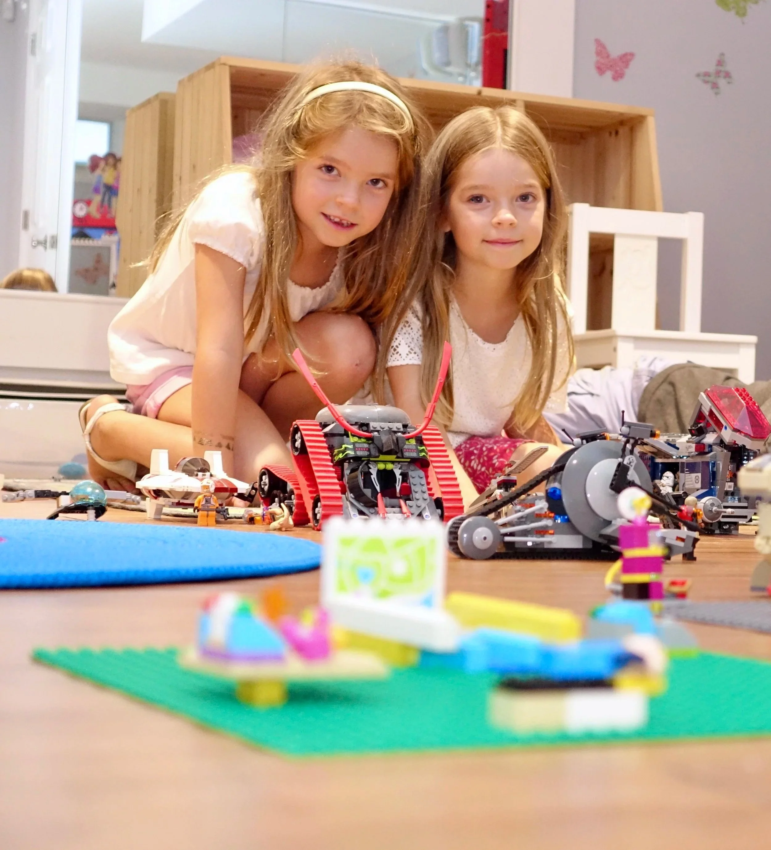 Two young girls with long hair posing next to a table full of Lego robots and toys, in a room with colorful decorations and a mirror in the background.