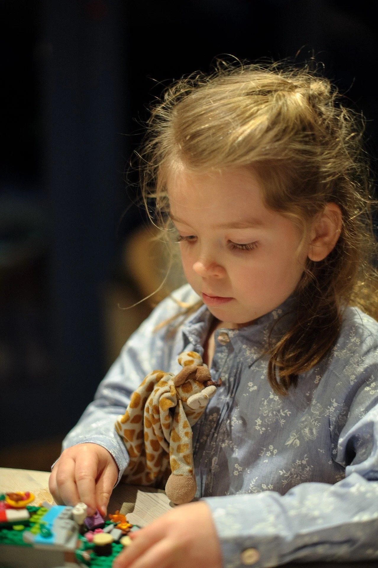Young Celine Samame playing with small colorful LEGO pieces on a table, holding a plush giraffe toy.