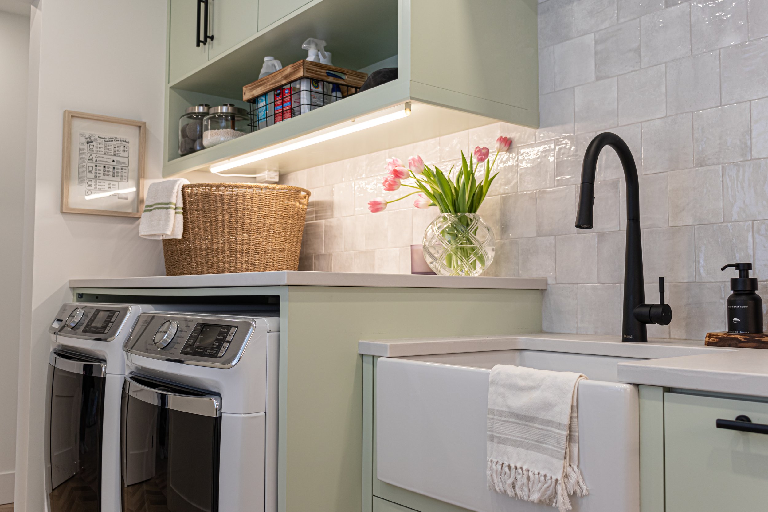 Laundry room with light green cabinets, a white countertop, a black faucet, a tiled backsplash, pink tulips in a glass vase, and a white farmhouse sink. There are two front-loading laundry machines below the counter and a woven basket on the counter. A framed chart hangs on the wall.