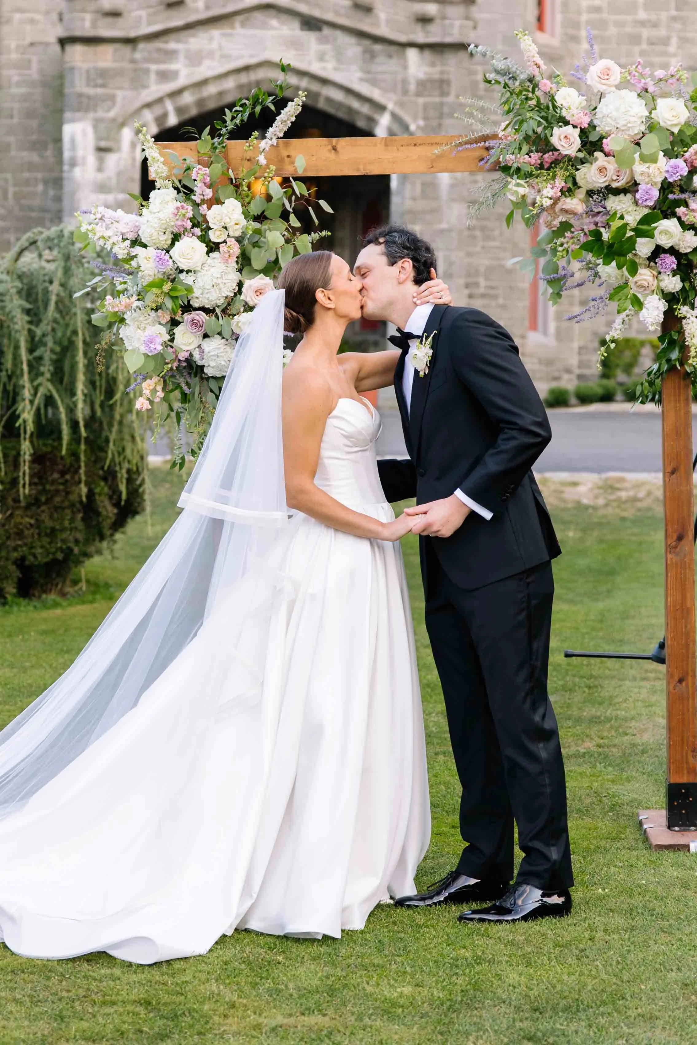 Bride and groom kissing under a floral arch at their outdoor wedding ceremony.